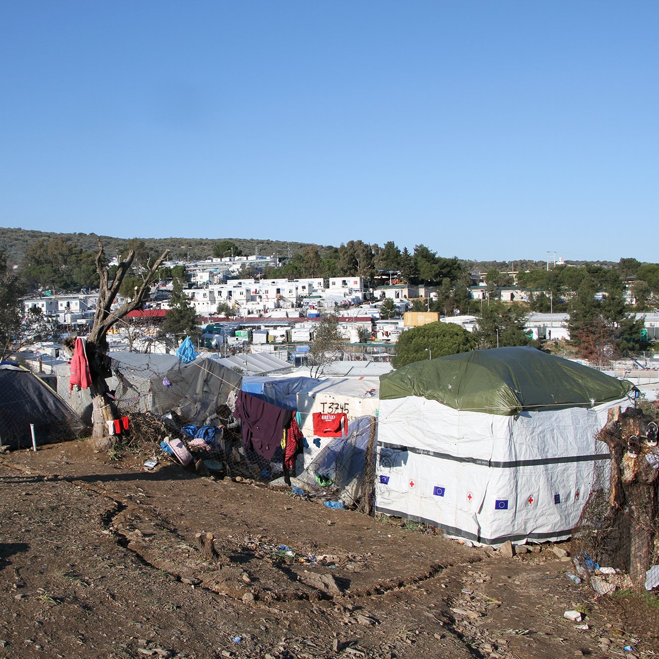 تصویر نشان‌دهنده یک منطقه با وضعیت نامناسب است که در آن چادرهایی به عنوان محل سکونت نصب شده‌اند. در پس‌زمینه، زمین‌های ناهموار و درختانی مشاهده می‌شود. پس‌زمینه شهری با ساختمان‌هایی دیده می‌شود که نسبتاً به چادرها نزدیک هستند. احساس کلی تصویر بیانگر شرایط سخت زندگی افرادی است که در این چادرها سکونت دارند. آسمان آبی و روز روشن است، اما وضعیت محیط نشان‌دهنده فقر و مشکلات معیشتی است.