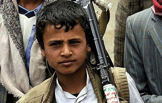 L'image montre un jeune garçon, probablement dans un contexte difficile. Il porte des vêtements simples et a un fusil en bandoulière. Son regard est sérieux, et il semble faire partie d'un groupe plus large, qui est à l'arrière-plan. Les vêtements des autres personnes indiquent un environnement rural ou de conflit. L'atmosphère générale de l'image témoigne d'un contexte de tension.