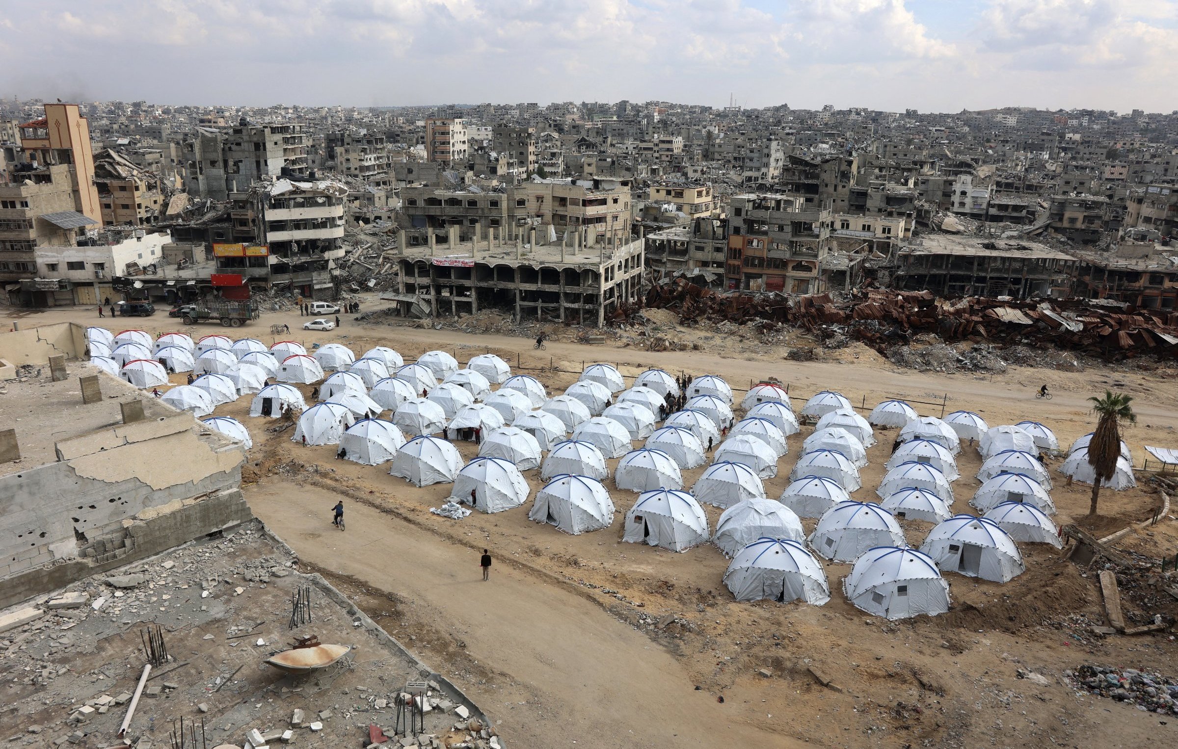 L'image montre une vaste zone de tentes blanches installées sur un terrain aride, entourée de bâtiments en ruines. Les tentes semblent former un camp, probablement pour des personnes déplacées ou des réfugiés. En arrière-plan, on peut voir des structures détruites, ce qui indique un environnement de conflit ou de catastrophe. Le ciel est nuageux, et l'atmosphère générale évoque la désolation et la nécessité d'abri.