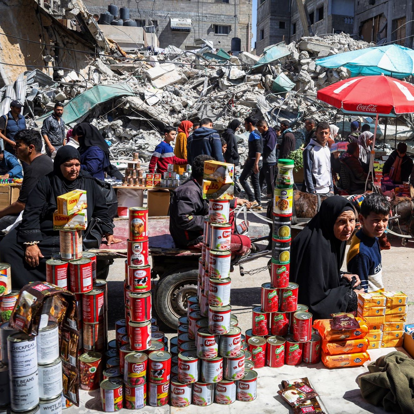 The image depicts a busy street scene in an area that appears to have experienced significant damage, as evidenced by the rubble and destroyed buildings in the background. In the foreground, a woman wearing a black hijab is sitting beside a young boy. They are surrounded by stacks of canned goods and other products that appear to be for sale, indicating a market or makeshift shop. People are seen walking through the street, some engaging in commerce amid the chaotic surroundings. Colorful umbrellas, likely providing shade, add contrast to the scene. Overall, it captures a moment of resilience and daily life amid challenging conditions.
