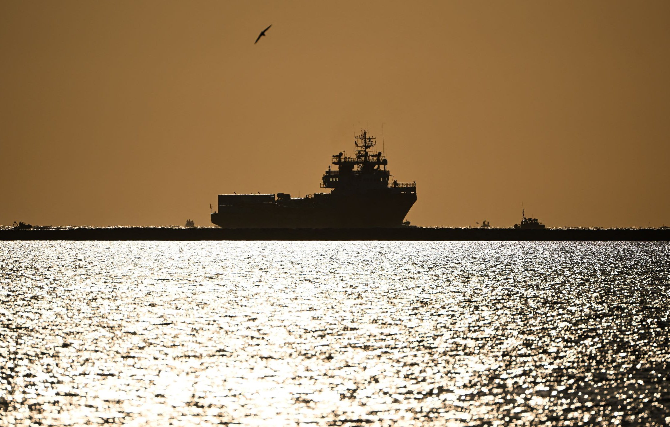 The image depicts a silhouette of a large ship on water during sunset or sunrise. The scene is bathed in a warm golden light, creating a dramatic contrast between the dark shape of the ship and the shimmering, reflective surface of the water. A seagull is seen flying in the sky, adding to the serene atmosphere of the moment. The horizon appears distant, enhancing the feeling of calmness and stillness in the scene.