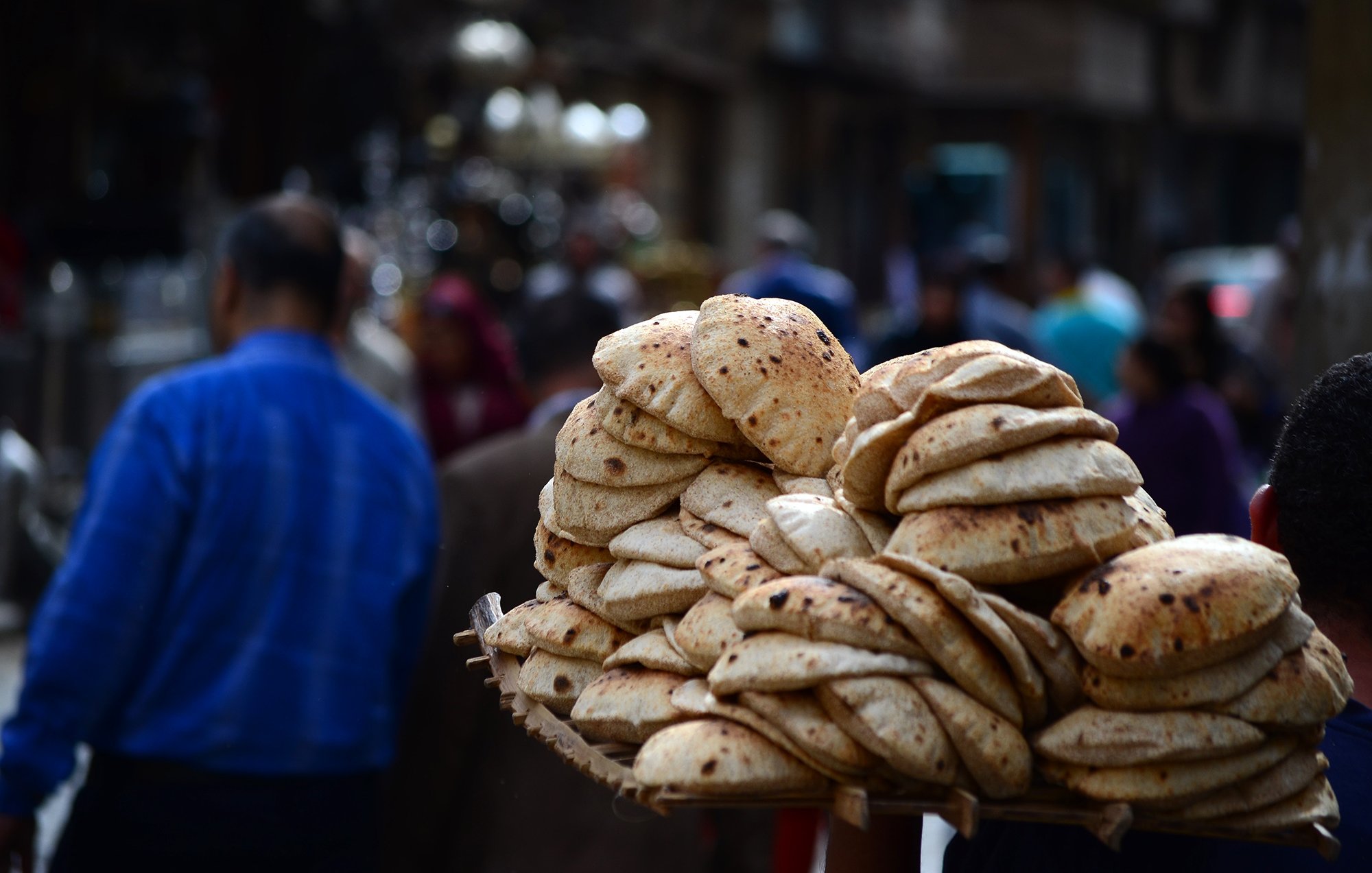 L'image montre un vendeur de pain dans une rue animée. Il porte une grande corbeille chargée d'un tas de pains ronds et dorés, souvent appelés "pita" ou "naan". En arrière-plan, on aperçoit des silhouettes de personnes se déplaçant dans la rue, créant une atmosphère vivante et dynamique. L'environnement semble plutôt urbain, avec des bâtiments et d'autres vendeurs visibles au loin.