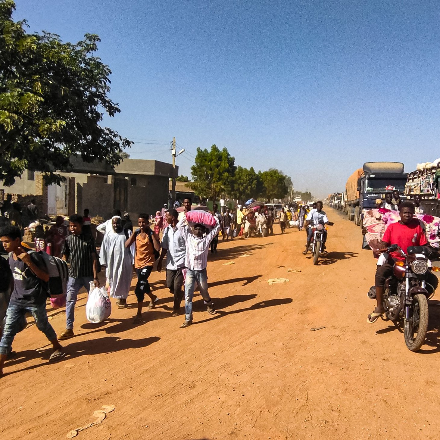 L'immagine mostra una scena di vita quotidiana lungo una strada in un ambiente urbano o semi-urbano. Si notano molte persone che camminano lungo il lato della strada, alcune con sacchi o beni in mano, e altre che sembrano discutere tra di loro. Sul lato opposto della strada ci sono veicoli in transito, tra cui camion e motociclette. La vegetazione circostante è presente, con alcuni alberi che offrono ombra. Il terreno appare polveroso, e l'atmosfera complessiva sembra vivace e movimentata.