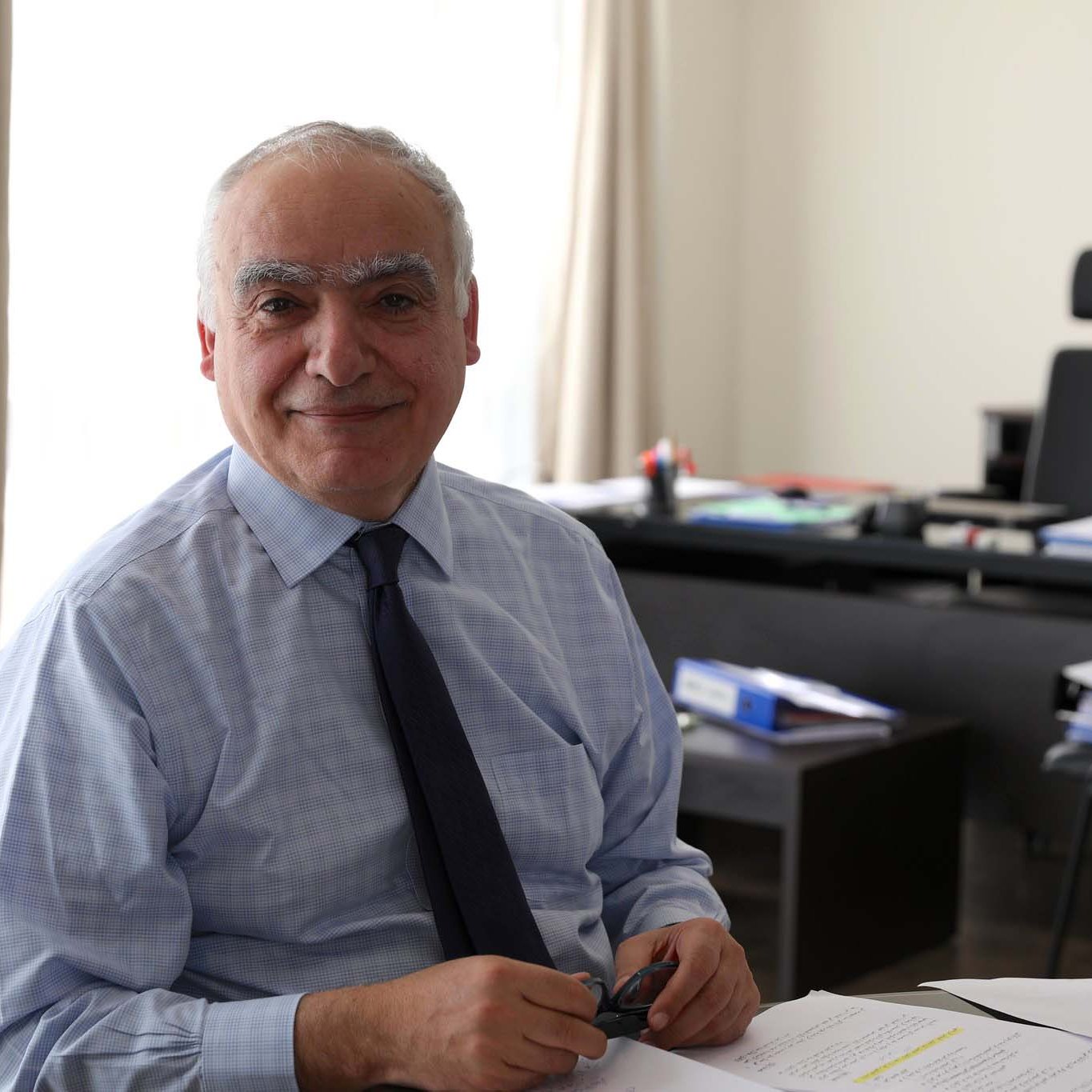 L'immagine mostra un uomo sorridente seduto a un tavolo in un ambiente di lavoro. Indossa una camicia e una cravatta, e sembra essere concentrato su alcune carte e documenti posti davanti a lui. Sullo sfondo, si intravedono elementi di un ufficio, come una scrivania con vari materiali e un ambiente luminoso grazie alla luce naturale che entra dalla finestra.