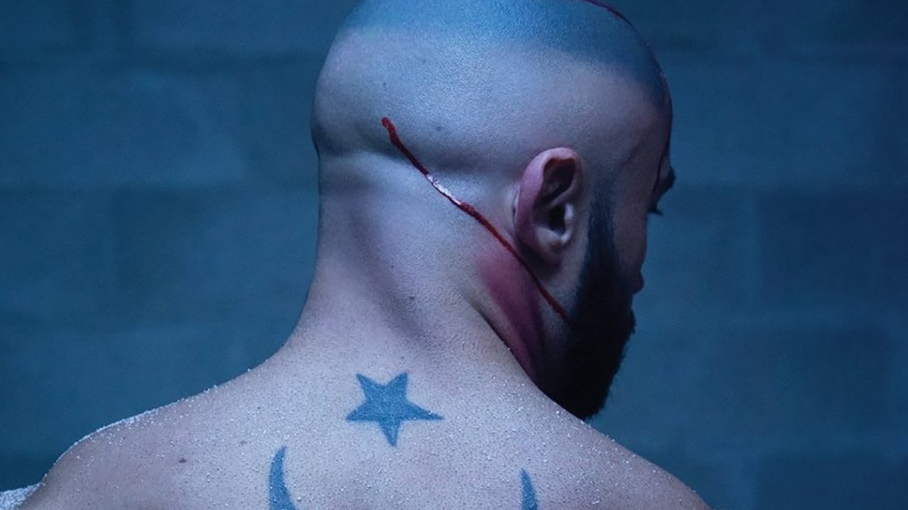 The image depicts a muscular, bald man from the back, showcasing a prominent tattoo on his back. The tattoo features a crescent moon and a star, which are often associated with various cultural and religious symbols. There are visible scars or cuts on his scalp and a streak of blood running down, suggesting a recent injury or intense situation. The background has a dark, moody atmosphere, possibly indicating a gritty or dramatic context.
