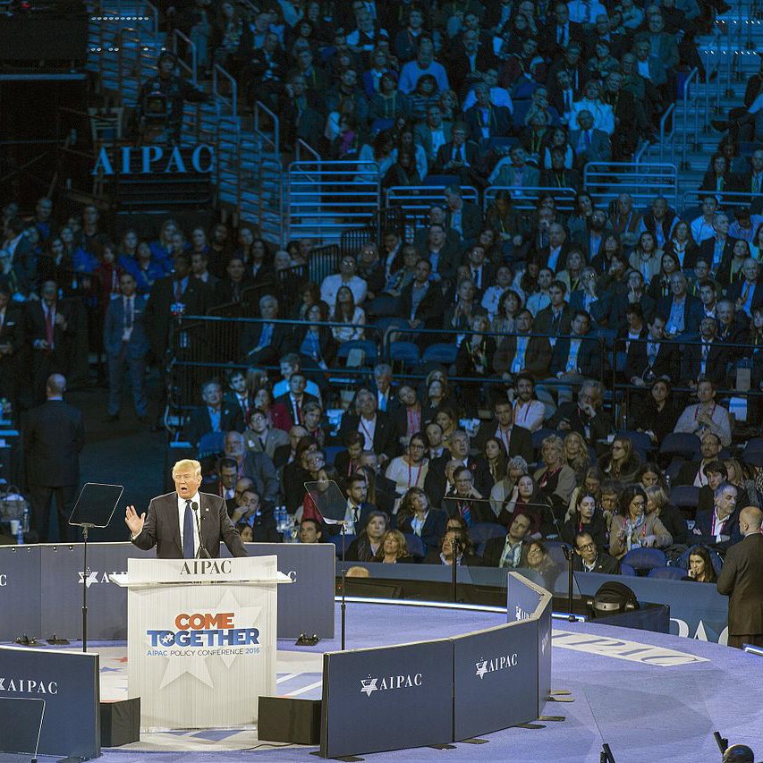 تظهر الصورة مشهداً لمؤتمر معبد مخصص لجمعية "AIPAC"، حيث يتحدث متحدث على المنصة أمام جمهور كبير. المنصة محاطة بأعلام وشعارات الجمعية، كما أن الإضاءة تلقي ظلالاً زرقاء على الحضور. يبدو أن المتحدث يناقش قضية مهمة، حيث يبدو الحضور متفاعلاً مع حديثه.