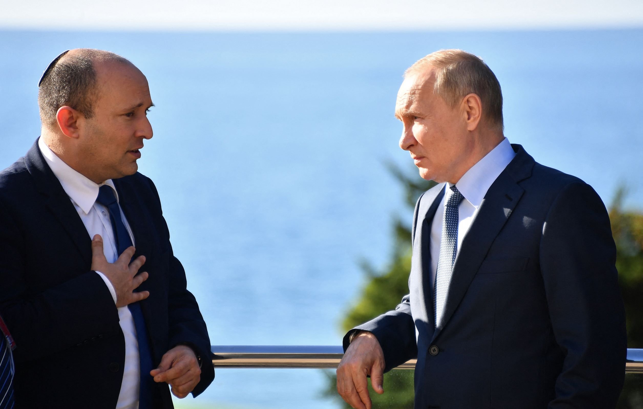 The image shows two men engaged in conversation by a seaside. One man is wearing a suit and has some form of head covering, indicating he may be of Jewish faith. The other man is also dressed in a suit and appears to be listening intently. They are both standing outdoors with a view of the water in the background. The setting suggests a formal or diplomatic meeting.