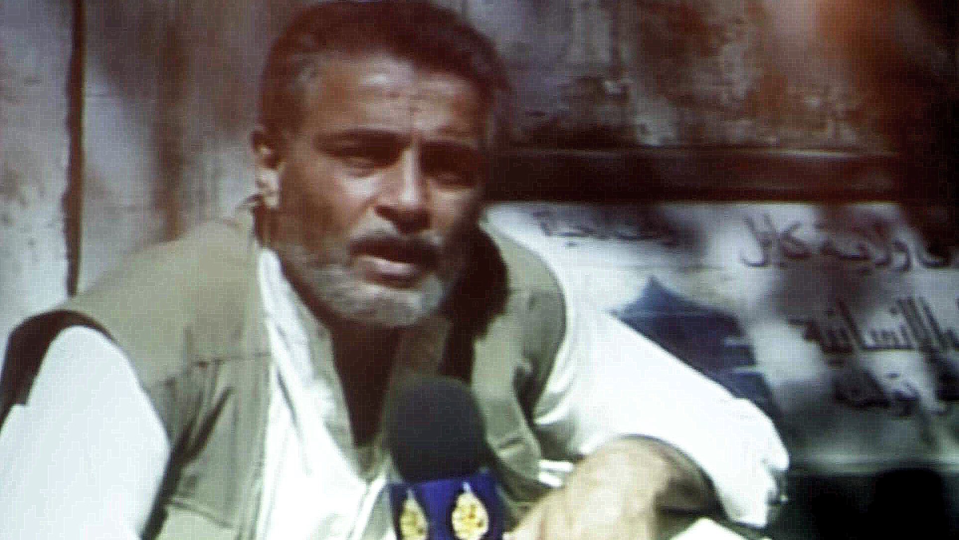 L'image montre un homme assis, tenant un micro et portant une tenue traditionnelle. Il a une barbe grise et semble parler avec intensité, probablement en train de faire un reportage. En arrière-plan, il y a un panneau ou une affiche qui n'est pas clairement visible. L'éclairage de l'image semble un peu flou, ajoutant une ambiance particulière à la scène.