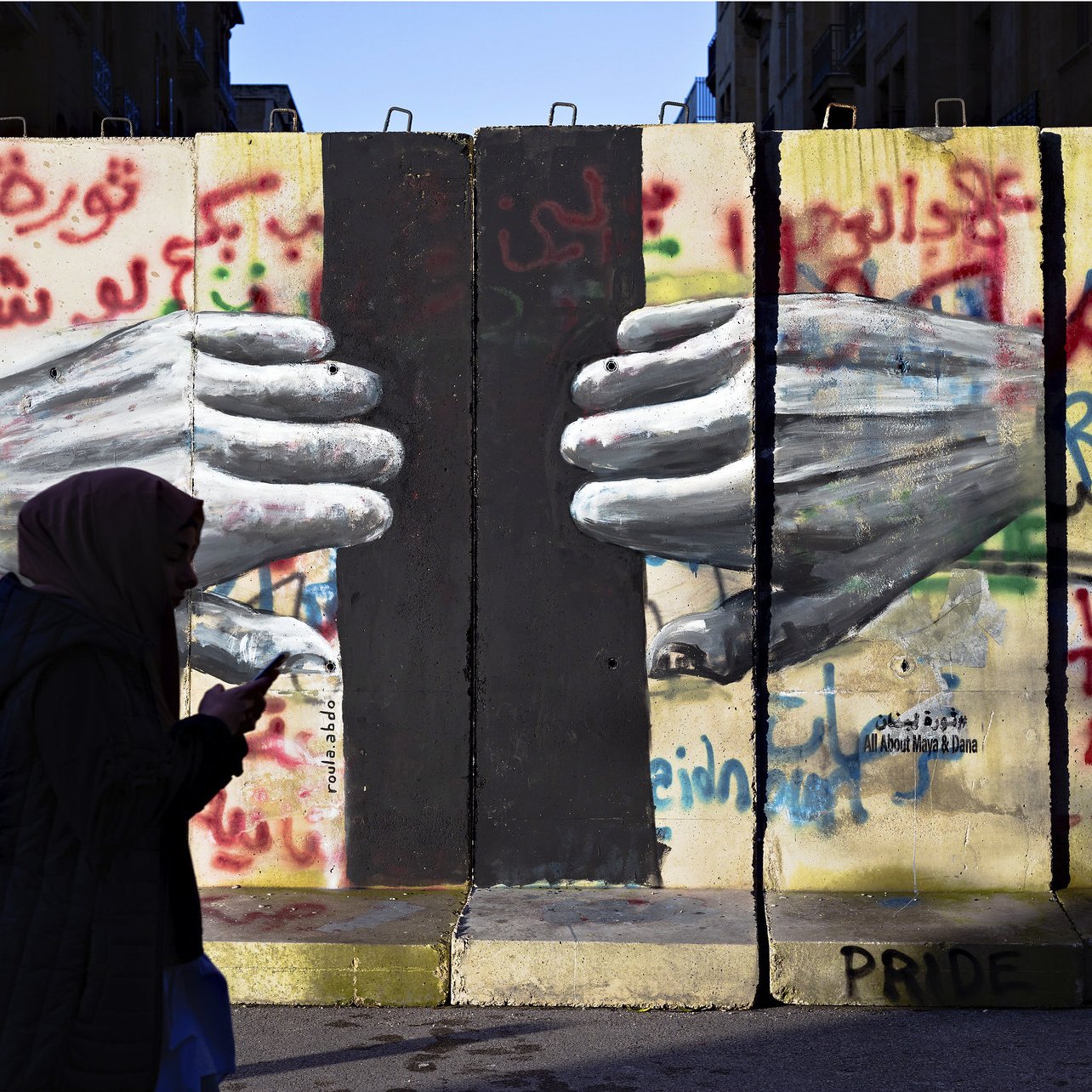 L'image montre un mur avec des graffitis colorés, dont le mot "Révolte" et d'autres messages en arabe et en anglais. Au centre, on peut voir des mains peintes qui semblent se toucher à travers une fissure du mur. Une silhouette d'une personne portant un hijab marche devant ce mur, avec un téléphone à la main. L'ensemble évoque des thèmes de résistance et de lutte sociale.