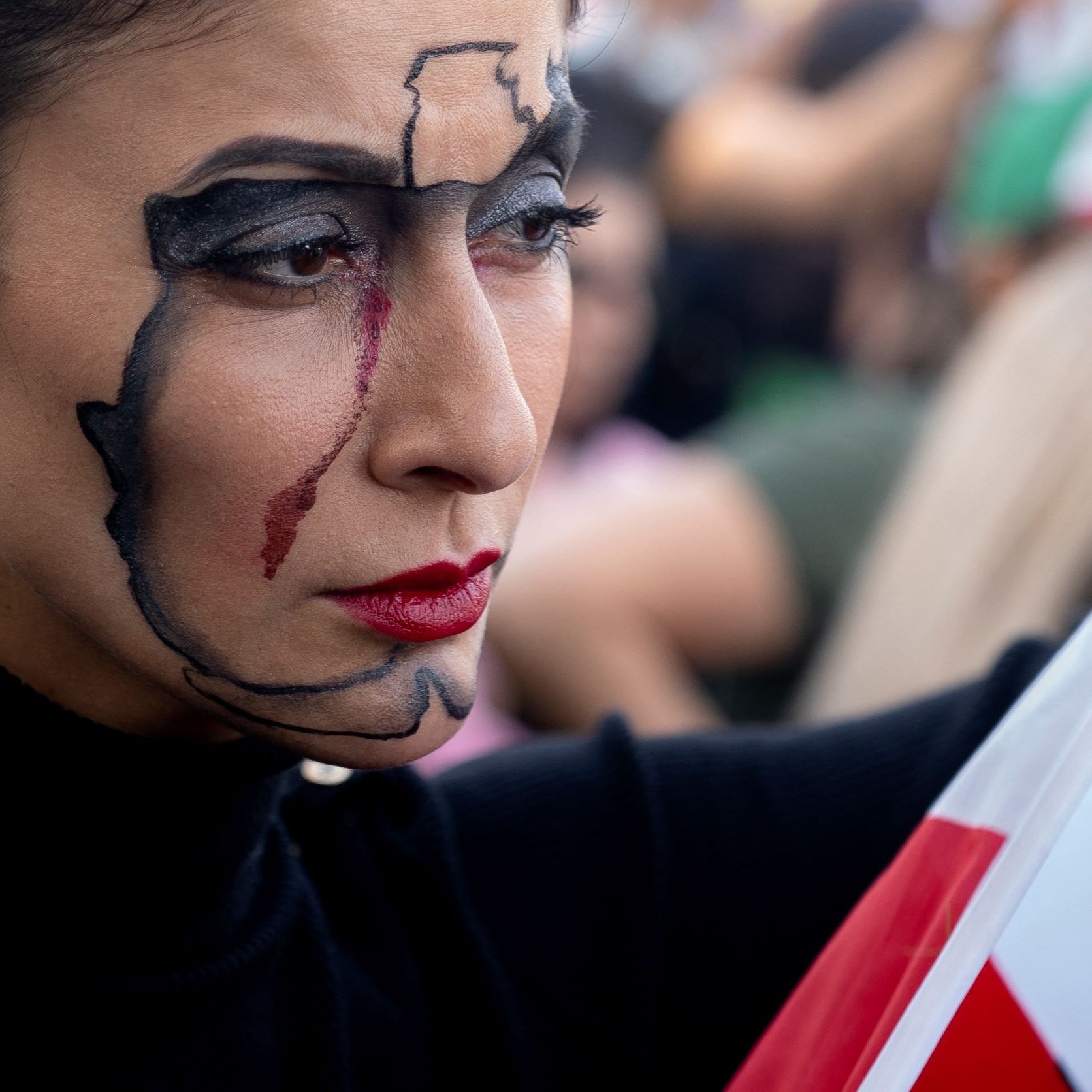 En la imagen se puede ver a una mujer con un maquillaje artístico que representa un mapa de México en su rostro, con un trazo destacado en negro y una lágrima roja que corre por su mejilla. Ella sostiene una bandera de México, que se encuentra en primer plano, mientras que en el fondo se vislumbran otras personas y un ambiente de manifestación. Su expresión es seria y reflexiva, lo que sugiere un fuerte mensaje emocional o político.