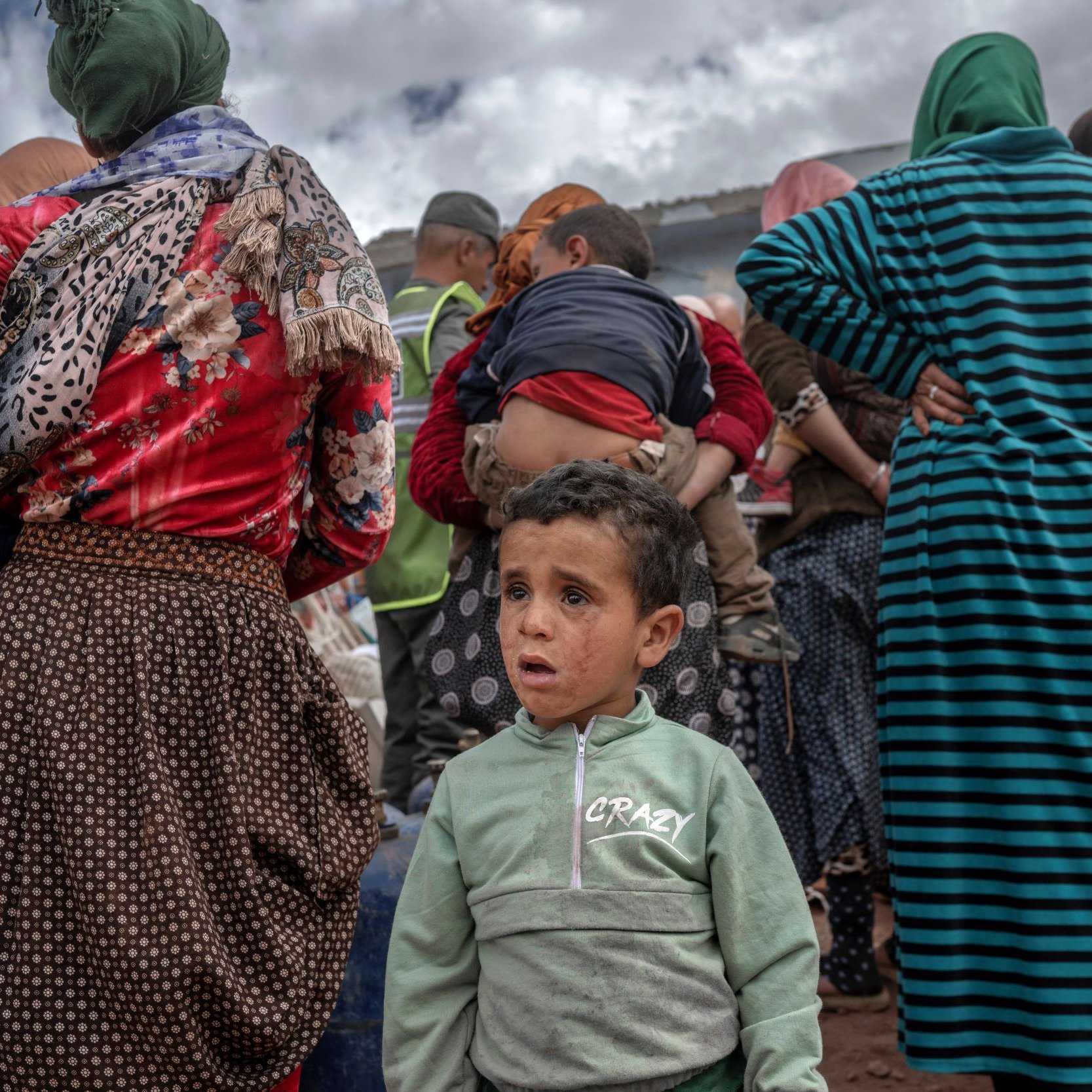La imagen muestra a un grupo de personas de espaldas, predominantemente mujeres, que parecen estar en un ambiente rural. La ropa que llevan es variada, con patrones y colores diversos. En el primer plano, un niño pequeño aparece al frente, mirando hacia la cámara con una expresión que podría reflejar confusión o preocupación. El fondo sugiere un cielo nublado y la escena evoca una sensación de comunidad y un posible evento o reunión.