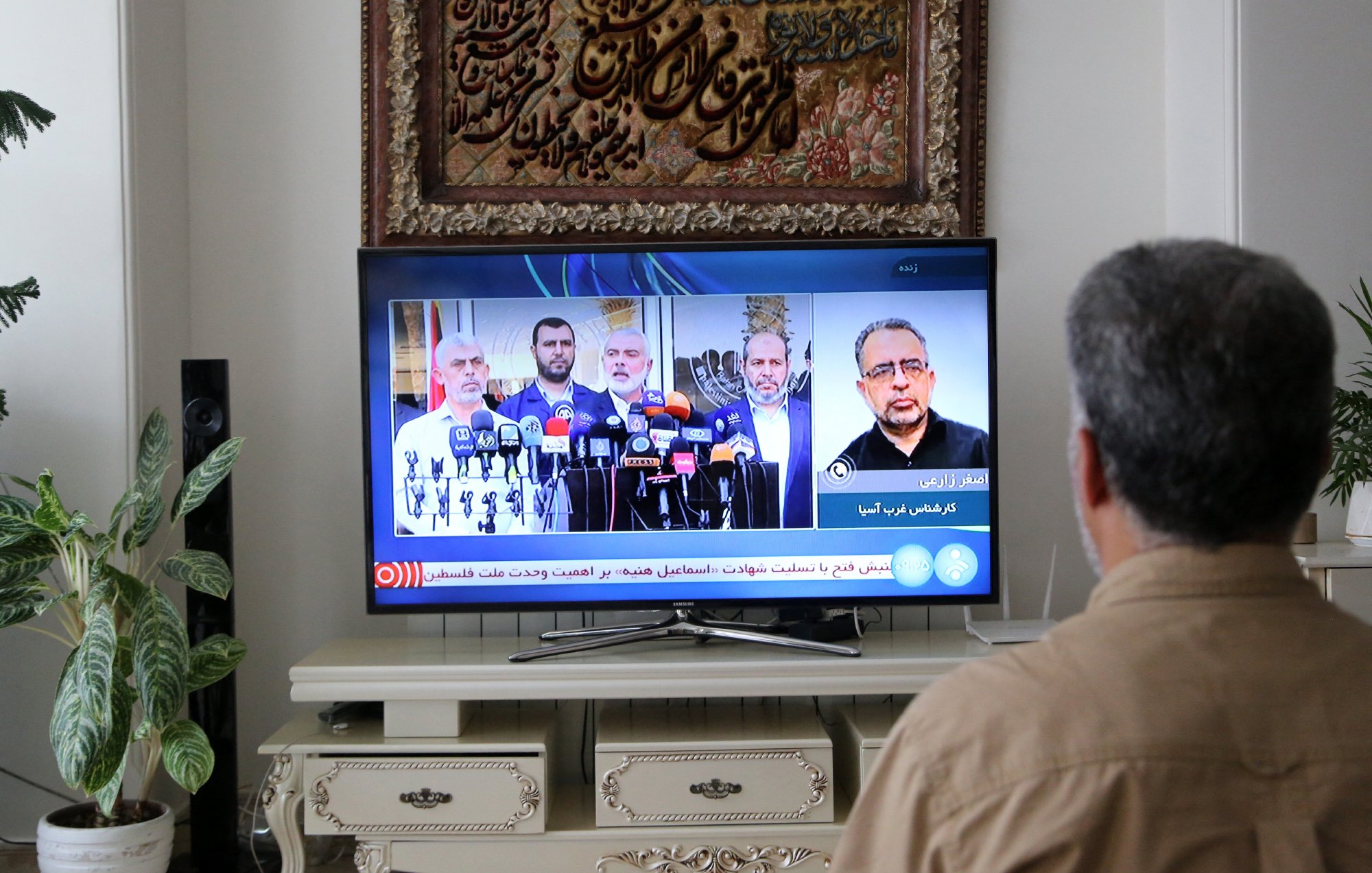 A person is sitting on a couch, facing a television that is displaying a news broadcast featuring several speakers at a press conference. In the background, there is a decorative wall hanging and a plant to the side.
