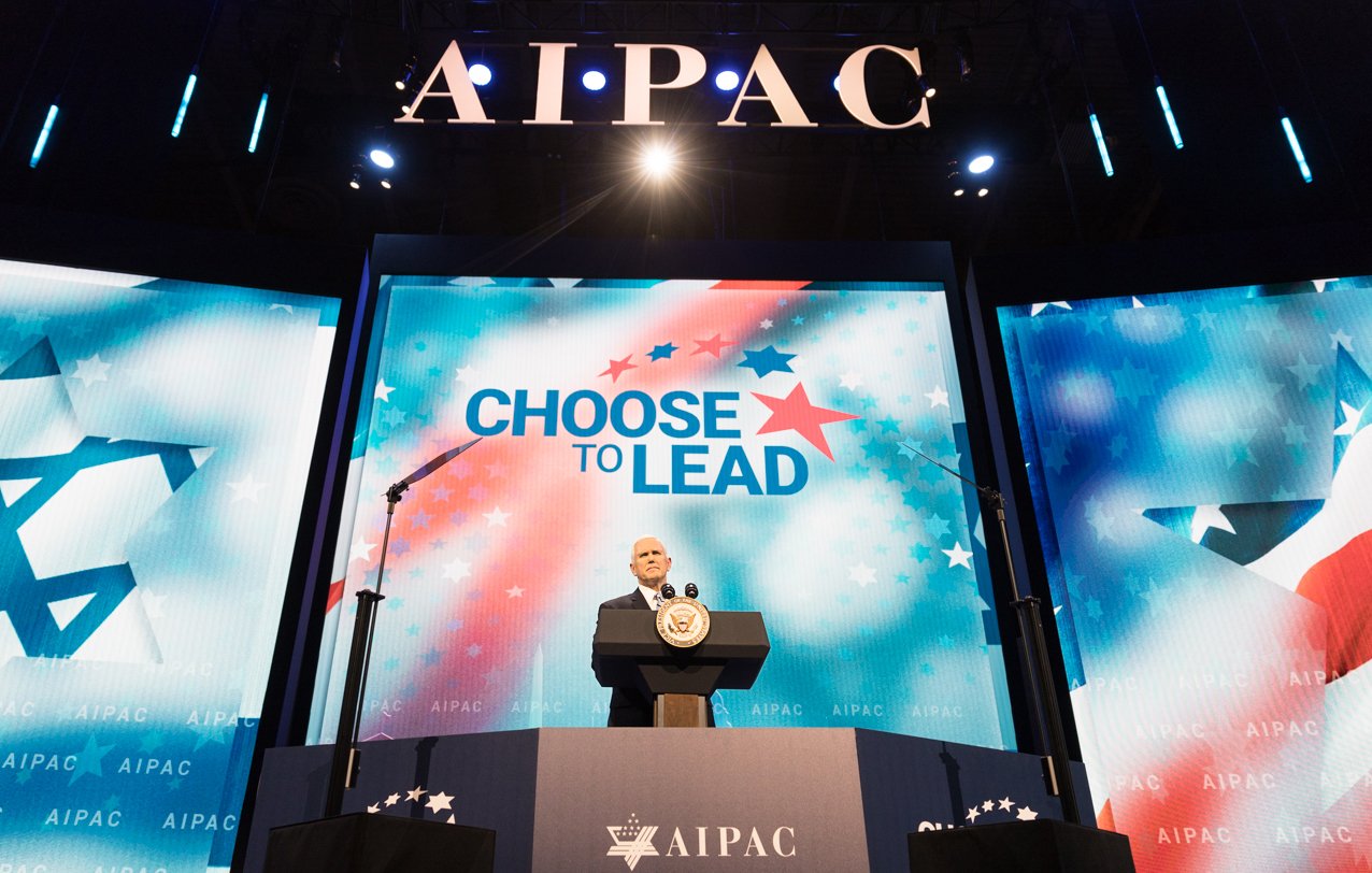 تظهر الصورة منصة خطابة كبيرة تحمل شعار "AIPAC" في الخلفية. يتواجد متحدث خلف ميكروفون على المنصة، مع وجود شاشات عرض كبيرة على الجانبين تظهر عبارة "اختَر القيادة" مع نجوم وشعارات ذات طابع إسرائيلي. الإضاءة تركز على المتحدث، مما يبرز أهمية الحدث الذي يُعقد.