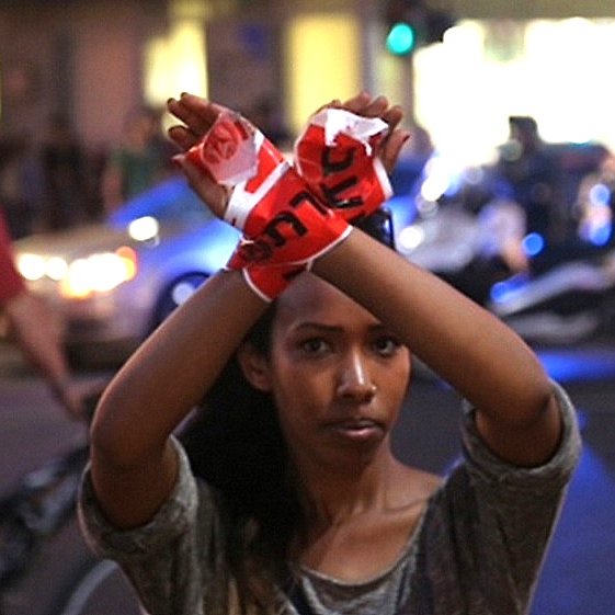 الصورة تظهر شابة ترفع يديها بطريقة تعبر عن الاحتجاج أو الاعتصام، حيث يتم ربط يديها بشريط يحمل كلمة أو شعار معين. الخلفية تحتوي على مشاهد حضرية، مع وجود أشخاص آخرين وسيارات تضيء بالأنوار. الأجواء تعكس لحظة من التوتر أو الحماسة، مما يدل على وجود حدث احتجاجي أو اجتماعي.