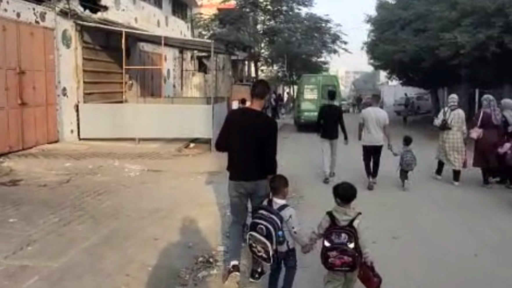 Un uomo cammina con due bambini, mentre altre persone si muovono in una strada cittadina.