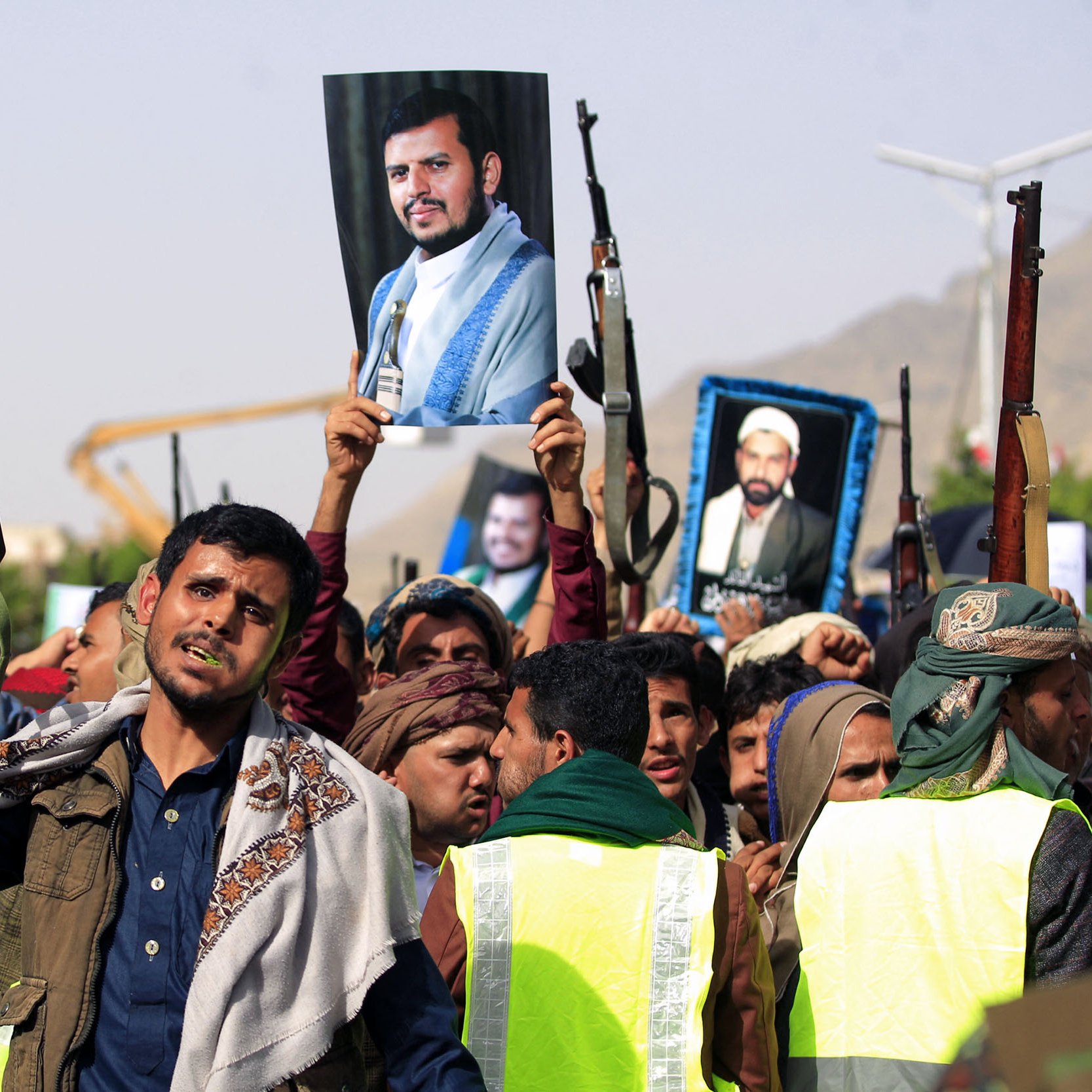 در این تصویر، گروهی از مردم در حال برگزاری یک تجمع هستند. تعدادی از شرکت‌کنندگان سلاح‌های خود را بلند کرده‌اند و تعدادی دیگر تصاویری از یک فرد را در دست دارند. این تجمع به نظر می‌رسد که از جنبه سیاسی و اجتماعی برخوردار باشد. مردمی که در تصویر دیده می‌شوند، لباس‌های سنتی و محلی به تن دارند و برخی از آن‌ها روسری‌هایی به سر بسته‌اند. پس‌زمینه تصویر شامل تعدادی سازه و فضای باز است.