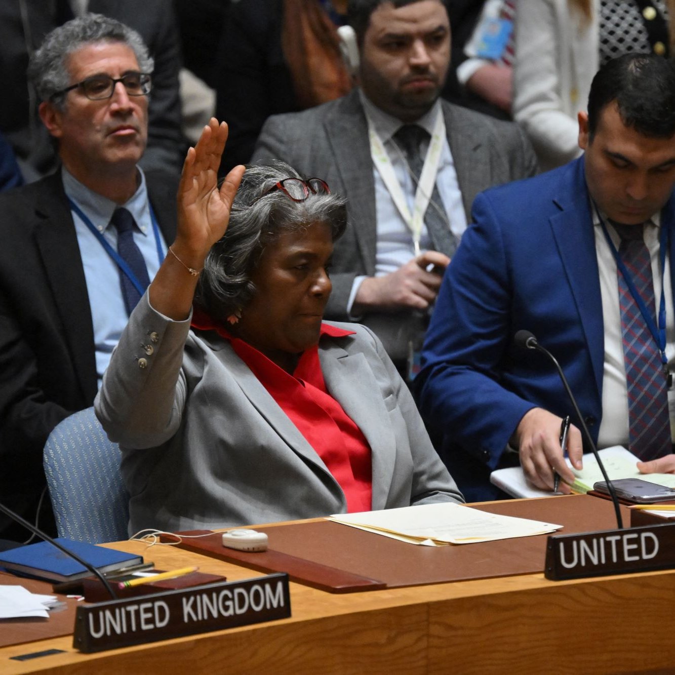 L'immagine mostra un incontro del Consiglio di Sicurezza delle Nazioni Unite. Una donna, che sta al centro della scena, alza la mano, apparentemente per esprimere un'opinione o richiedere la parola. Indossa un blazer grigio con una camicia rossa e sembra essere in fase di comunicazione. Attorno a lei, ci sono diversi rappresentanti di altri paesi, tra cui stati come Regno Unito e altri, seduti con atteggiamenti attenti. L'atmosfera sembra seria e formale, tipica di una riunione diplomatica.