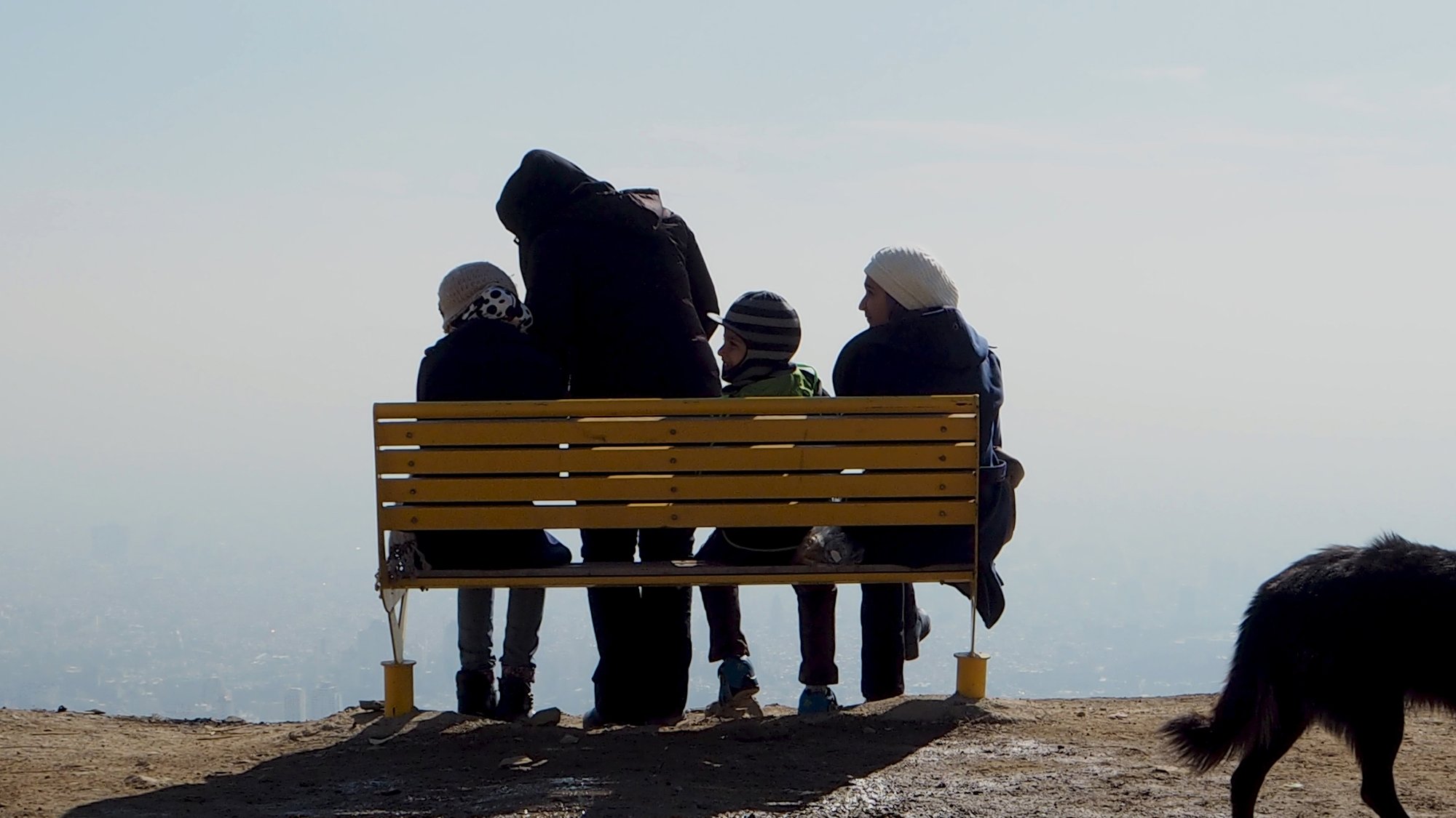 L'image montre un groupe de quatre personnes assises sur un banc jaune sur une colline ou un sommet. Ils sont tournés vers un paysage brumeux, peut-être en admirant la vue. Parmi eux, on peut voir un adulte et trois enfants. L'adulte porte un manteau sombre tandis que les enfants portent des bonnets. À droite de l'image, un chien noir se tient sur le sol, se dirigeant vers l'extérieur du cadre. L'atmosphère semble tranquille et contemplative, avec une faible visibilité en raison de la brume.