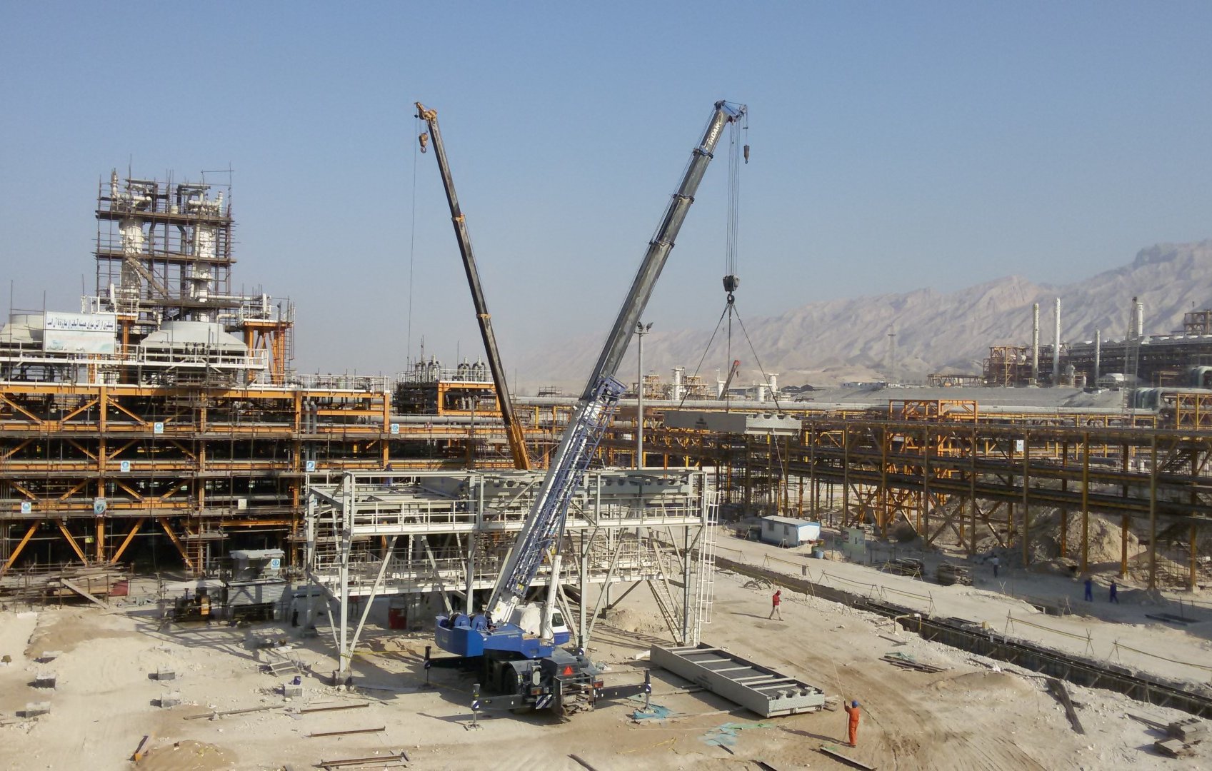 L'image montre un chantier de construction avec plusieurs grues sur le site. On peut voir des structures métalliques en voie d'achèvement à l'arrière-plan, ainsi que des véhicules de chantier et des ouvriers sur le site. Le paysage environnant est montagneux, et le ciel est dégagé, ce qui suggère une journée ensoleillée. Le chantier semble être à un stade de construction avancé, avec des matériaux disposés sur le sol.