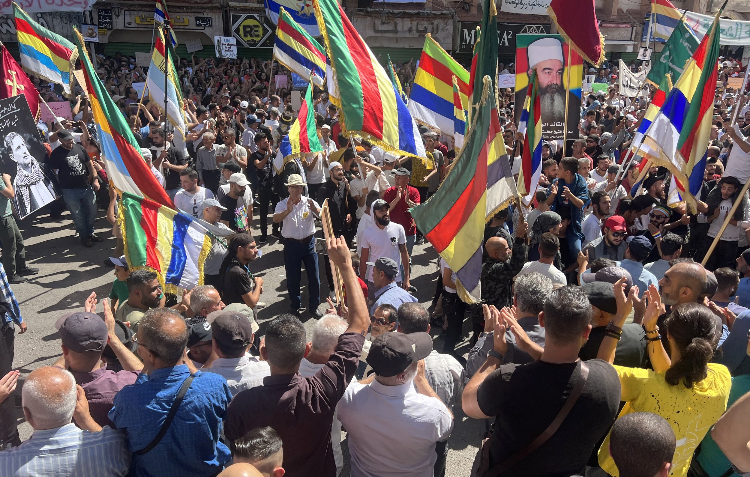 The image depicts a large crowd gathered in a public space, participating in a demonstration or protest. Many people are holding brightly colored flags that feature a mix of horizontal and vertical stripes. The atmosphere appears energetic and spirited, with participants actively engaging with one another. Some individuals are raising their hands and clapping, suggesting a celebratory or passionate mood. In the background, various signs and posters can be seen, indicating that the event is likely focused on a specific cause or issue.