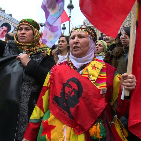 L'image montre une manifestation avec des personnes brandissant des drapeaux et des bannières. Certaines femmes portent des vêtements traditionnels et des accessoires colorés, et elles expriment leurs revendications avec des gestuelles passionnées. Il y a également des portraits visibles en arrière-plan, suggérant un hommage à des individus particuliers. L'ambiance semble être à la fois festive et engagée, avec une forte symbolique culturelle.