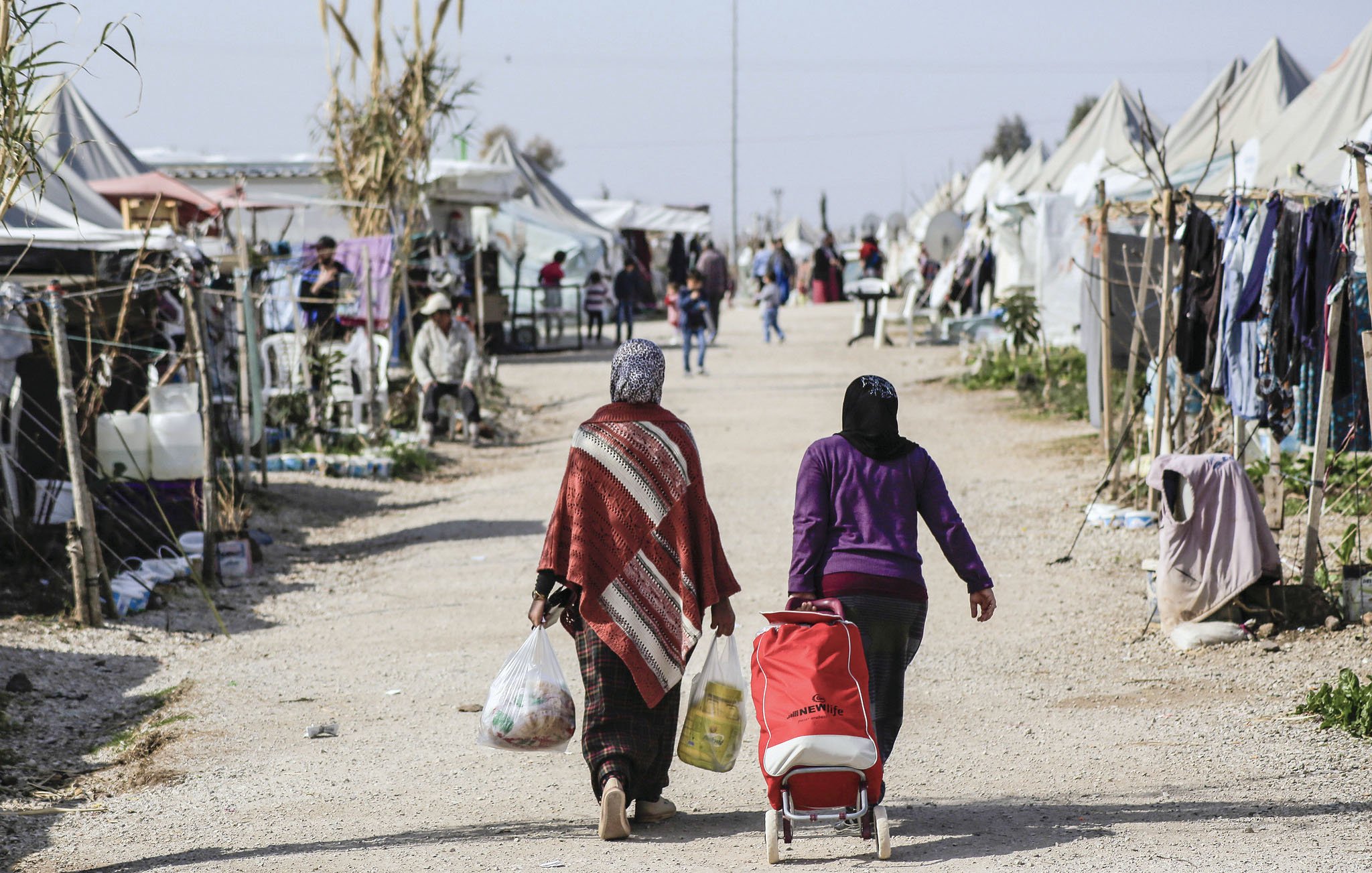 The image depicts two women walking down a dirt path in a makeshift camp. One woman carries bags of supplies, while the other pushes a stroller. On either side of the path, there are tents and structures that suggest a temporary living situation. The environment appears to be bustling with activity, as other individuals can be seen in the background, some seated or engaging in conversation. The scene conveys a sense of community within the challenges of a refugee or displaced persons camp.