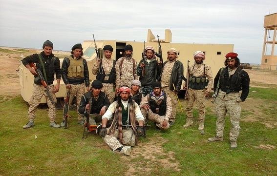 L'image montre un groupe de personnes en tenue militaire, armées et posant devant un véhicule. Ils semblent être sur un terrain en extérieur, peut-être dans un environnement désertique ou rural. Les hommes affichent diverses expressions et postures, certains sont debout tandis que d'autres sont assis. Leurs vêtements et accessoires suggèrent qu'ils font partie d'une unité militaire ou paramilitaire. En arrière-plan, on aperçoit un bâtiment et une tour d'eau.