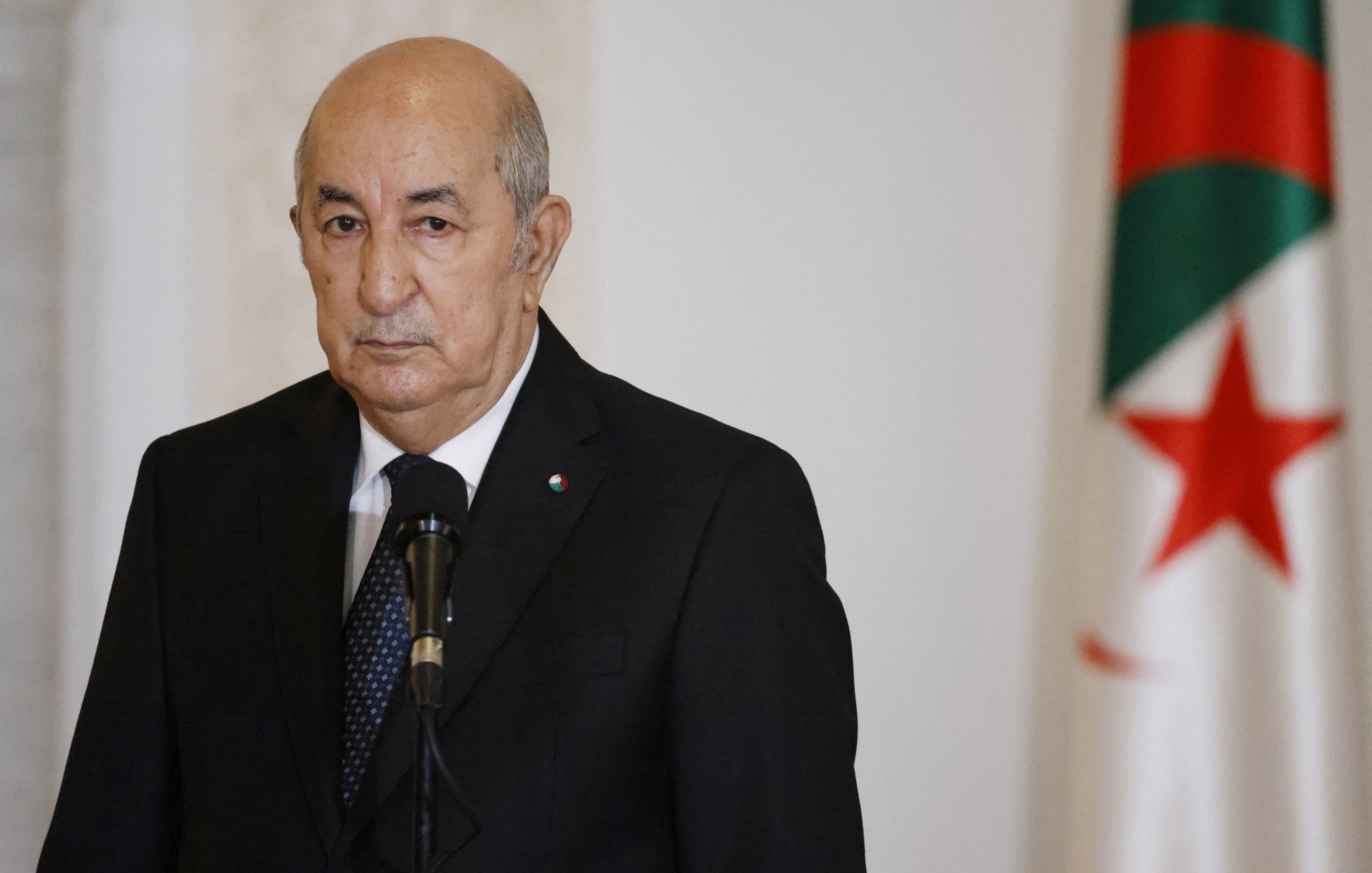 L'image montre un homme en costume sombre, regardant vers l'avant avec une expression sérieuse. Il se tient devant un microphone, et en arrière-plan, on peut voir un drapeau. Le drapeau présente des couleurs vertes et rouges avec une étoile et un croissant. L'ambiance de l'image semble solennelle, suggérant un contexte formel ou officiel.