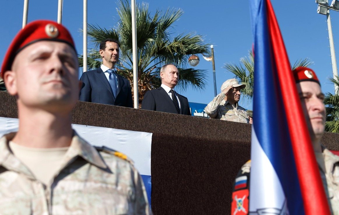 The image depicts a military ceremony or event, featuring several individuals of importance. In the foreground, soldiers in military uniforms with red berets are standing at attention. In the background, a group of officials is present, including two prominent figures positioned together: one in a suit and tie and the other in military attire. There's a large flag in the foreground, possibly representing a nation involved in the event. The setting appears outdoors, with palm trees visible, suggesting a warm climate. The overall atmosphere seems formal and significant, likely indicative of a diplomatic or military alliance.