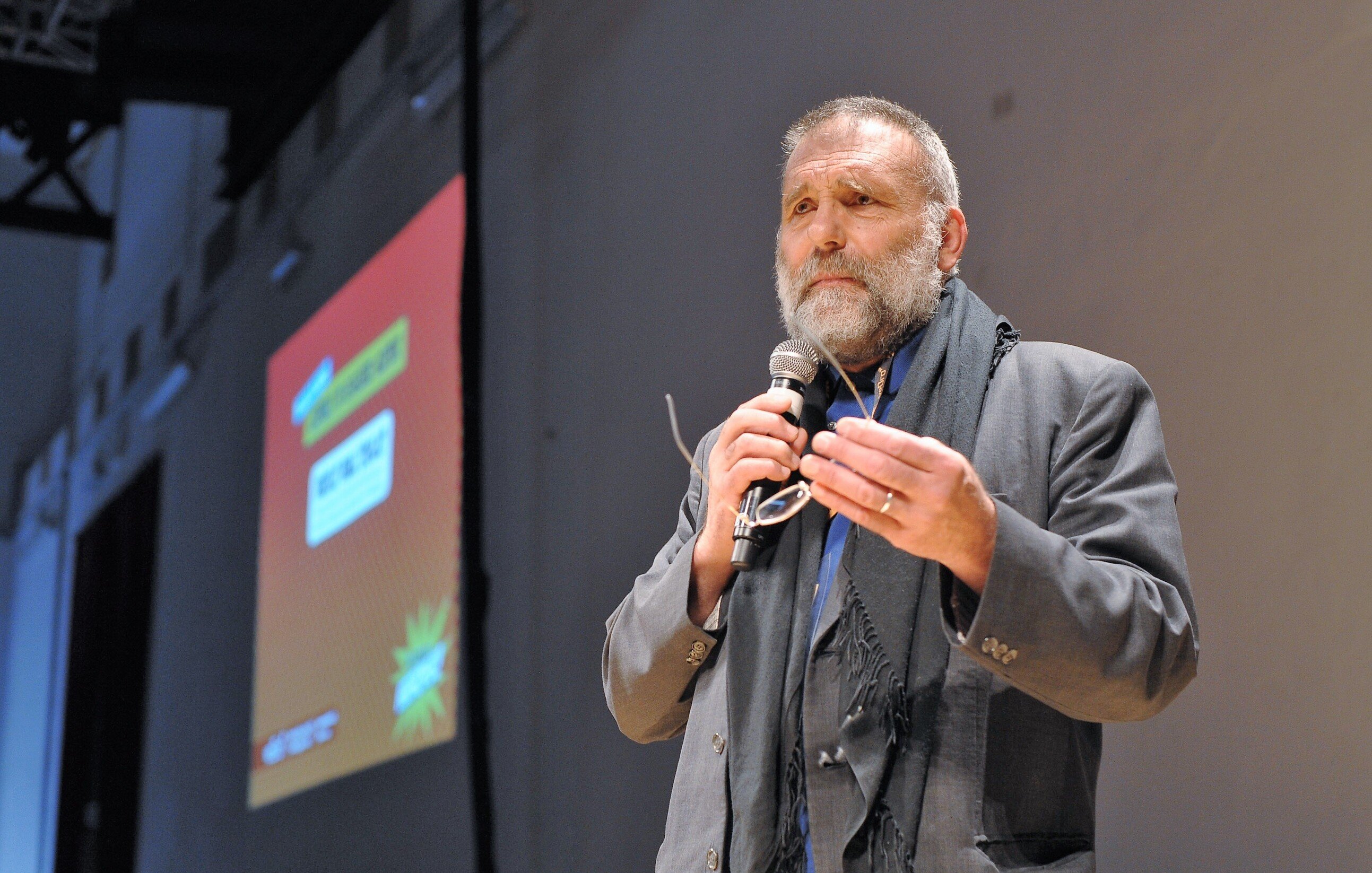 Nell'immagine si vede un uomo, probabilmente un relatore, che sta parlando al pubblico. Indossa un abito elegante e ha una sciarpa scura. Tiene un microfono in mano e sembra essere coinvolto in un discorso. Sullo sfondo c'è un grande schermo con delle grafiche colorate, ma il contenuto di ciò che viene mostrato non è visibile. L'atmosfera dell'immagine sembra essere formale e interattiva.