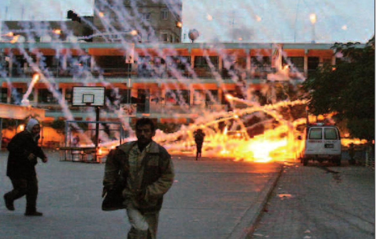 L'image montre une scène de chaos avec des explosions et des traînées de lumière dans le ciel. Des personnes semblent fuir dans une zone qui ressemble à une cour d'école, tandis qu'un bâtiment en arrière-plan est visible. L'environnement semble marqué par une tension intense, avec des traces d'incendie et des éclats lumineux qui suggèrent une situation dangereuse. L'atmosphère est sombre, renforçant le sentiment d'urgence et de désarroi.