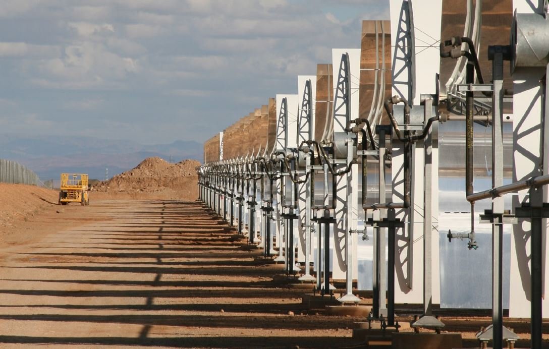 تظهر الصورة مجموعة من الألواح الشمسية المرتبطة بهياكل معدنية، تقع في منطقة صحراوية أو شبه صحراوية. الألواح مرتبة بشكل منتظم وموجهة نحو السماء، مما يشير إلى أنه يجري استغلال الطاقة الشمسية. في الخلفية، يمكن رؤية تلال ترابية وبعض السحب في السماء، مما يعطي انطباعًا عن مساحة واسعة ومفتوحة. تُظهر الصورة أيضًا طريقًا يسير فيه مركبة كبيرة تشير إلى وجود نشاط صناعي أو زراعي في المنطقة.