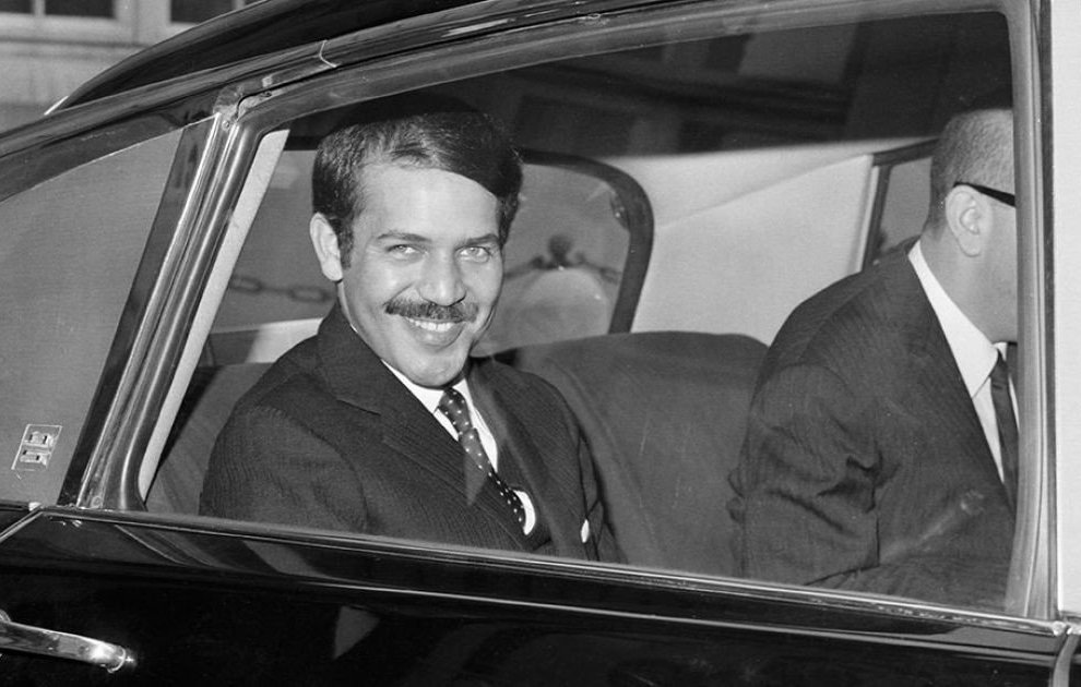 The image shows a man sitting in the back seat of a car, smiling at the camera. He is dressed in a formal suit and tie, with his hair neatly styled. Another person, partially visible, is seated next to him, also dressed in a suit. The setting appears to be a city, indicated by buildings in the background. The photograph has a classic black-and-white aesthetic, suggesting it was taken several decades ago.