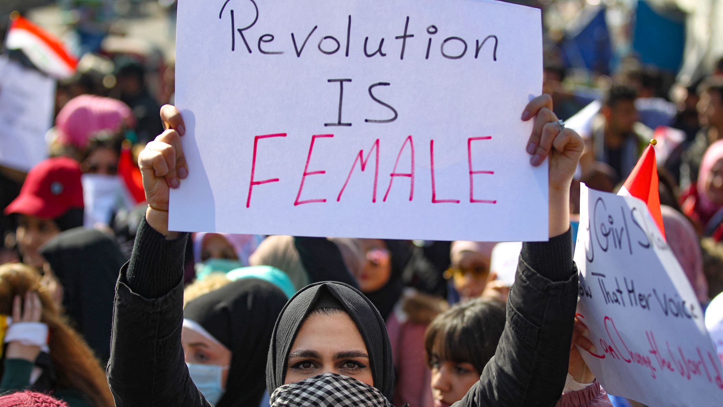 الصورة تظهر مجموعة من المتظاهرين في تجمع حاشد، حيث تحمل امرأة لافتة مكتوب عليها "الثورة أنثوية". يتواجد أفراد من مختلف الأعمار في الخلفية، كما يظهر بعضهم يرتدي الأوشحة والقبعات. الأجواء تعكس حماس المتظاهرين والتزامهم بقضية معينة.