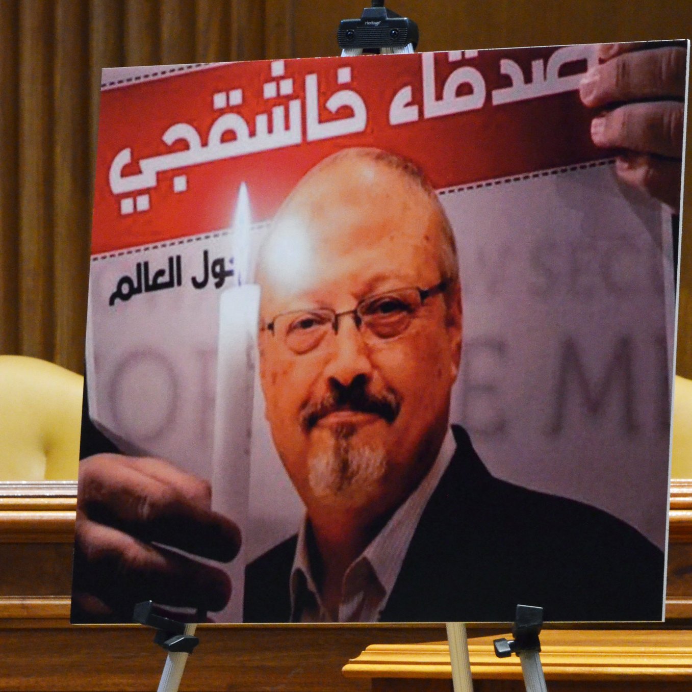 الصورة تظهر لوحة تحمل صورة لصحفي وهو يحمل شمعة مضاءة. خلفه يوجد نص عربي يرد فيه اسم "جمال خاشقجي". يبدو أن اللوحة تم استخدامها في مناسبة لتسليط الضوء على قضيته أو إحياء ذكراه، حيث يعتلي الصورة تعبير عن التضامن أو الاحتجاج. في الخلفية، هناك إعداد رسمي مع مقاعد فارغة.