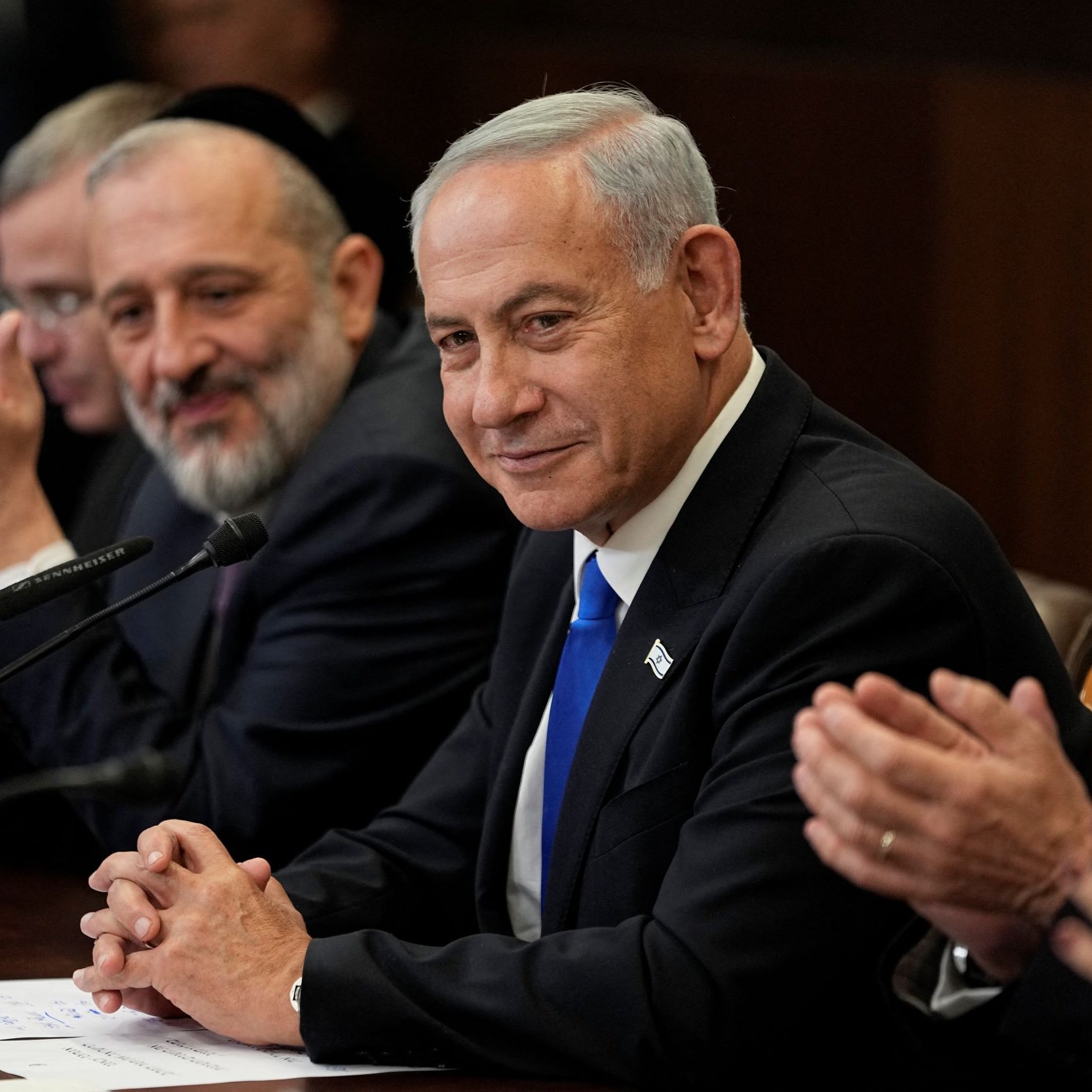 The image depicts a group of men seated around a table during a formal meeting. In the foreground, a man in a suit with a blue tie appears to be the focal point, wearing a slight smile. He is flanked by other men who are also dressed in formal attire. One man, likely a religious figure, is seen smiling broadly. The setting appears to be an official governmental or diplomatic environment, indicated by the wooden table and the serious demeanor of those present. There are papers and a microphone in front of them, suggesting they are engaged in a discussion or announcement.
