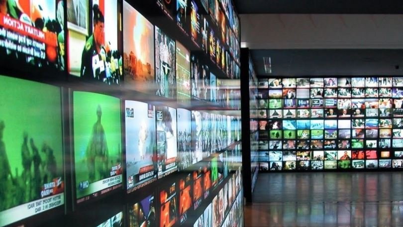 تظهر الصورة مجموعة كبيرة من الشاشات التلفزيونية المعلقة على حائط، وكل شاشة تعرض محتوى مختلف. تتنوع الصور التي تظهر على الشاشات بين الأخبار، والفيديوهات المتنوعة، مما يعطي انطباعًا عن مكان مفعم بالمعلومات والمرئيات. الألوان في الصورة تتراوح بين الأخضر والأحمر والأزرق، مما يخلق جوًا ديناميكيًا ومثيرًا للاهتمام. يُمكن أن تعكس الصورة فكرة الاستهلاك البصري الكبير للمعلومات في العصر الحديث.