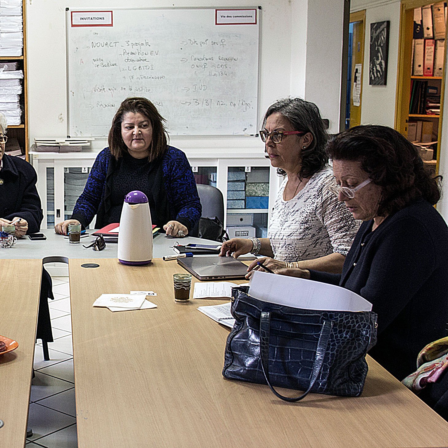 في الصورة، نرى مجموعة من النساء يجلسن حول طاولة في غرفة تحتوي على رفوف مملوءة بالأوراق والمستندات. يظهر على الطاولة بعض الأكواب والأطعمة الخفيفة، بينما يبدو أن المشاركات في اجتماع أو جلسة مناقشة. النساء يبدين مهتمات بما يحدث، حيثيزن التفاعل مع بعضهن البعض. الغرفة تملك أجواء مهنية، مع جدران بيضاء وسبورة خلفهن.