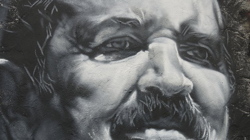 L'image représente une œuvre murale en noir et blanc, mettant en avant un visage masculin avec des traits expressifs. La personne a une moustache proéminente, et son expression semble montrer une certaine intensité. Les détails des ombres et des lumières apportent de la profondeur au portrait, accentuant les émotions qui s'en dégagent. Les contours sont soigneusement travaillés, ce qui donne une apparence réaliste au personnage.
