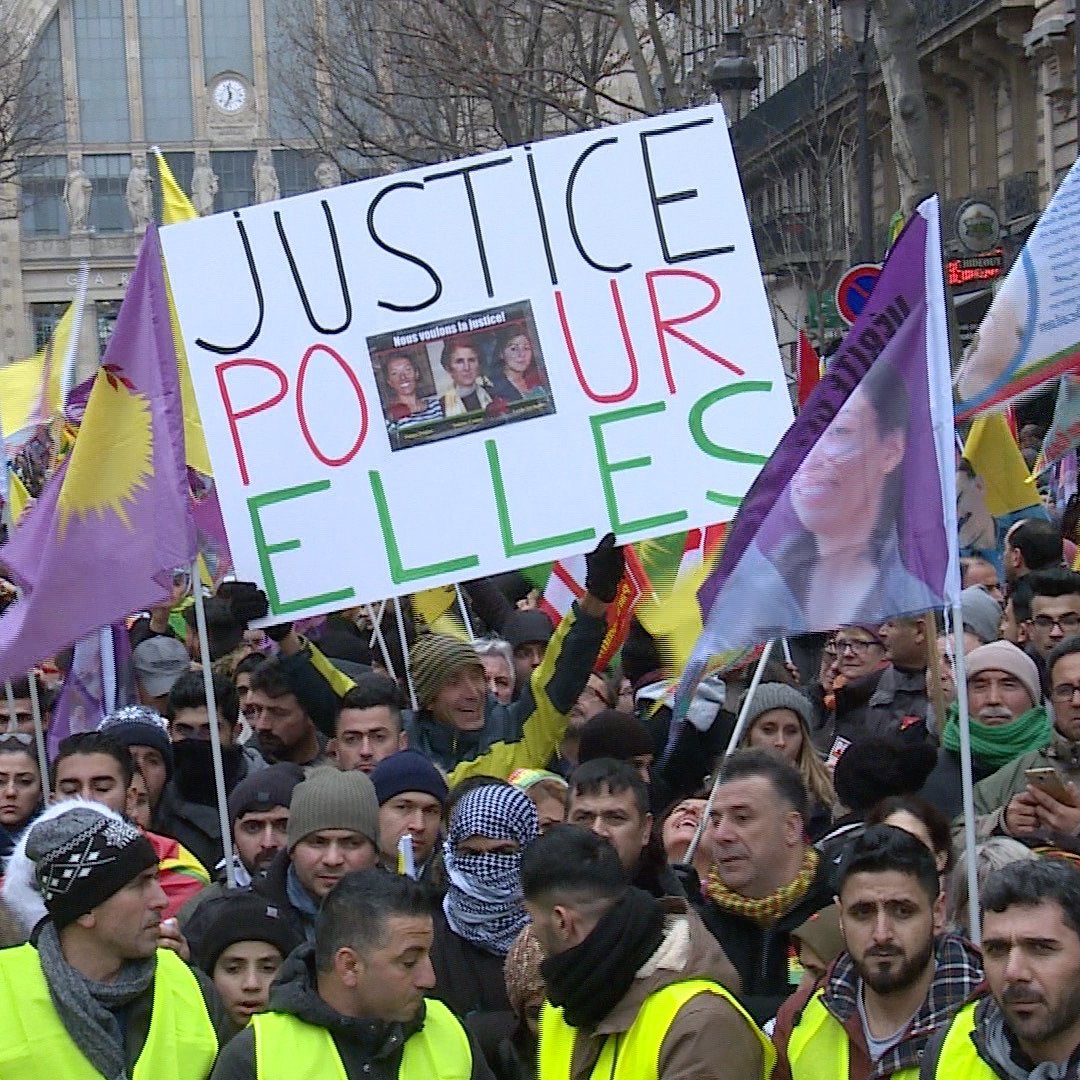 L'image montre une manifestation avec de nombreuses personnes rassemblées. Les manifestants portent des drapeaux colorés et des panneaux. L'un des panneaux affiche le message "JUSTICE POUR ELLES", suggérant un appel à la justice, probablement pour des femmes. Les visages des manifestants sont déterminés, et l'atmosphère semble impliquée et engagée.