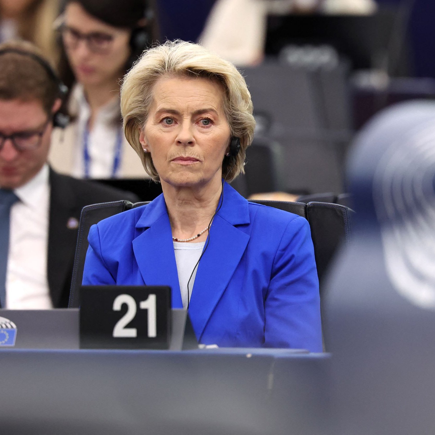 Nell'immagine si vede una donna seduta in un ambiente di tipo istituzionale, probabilmente durante una sessione del Parlamento Europeo. Indossa un blazer blu e ha i capelli biondi. Sembra concentrata e seria, mentre intorno a lei ci sono altre persone che ascoltano attentamente, alcune con cuffie. L'ambientazione è moderna, con posti a sedere e schermi visibili sullo sfondo.