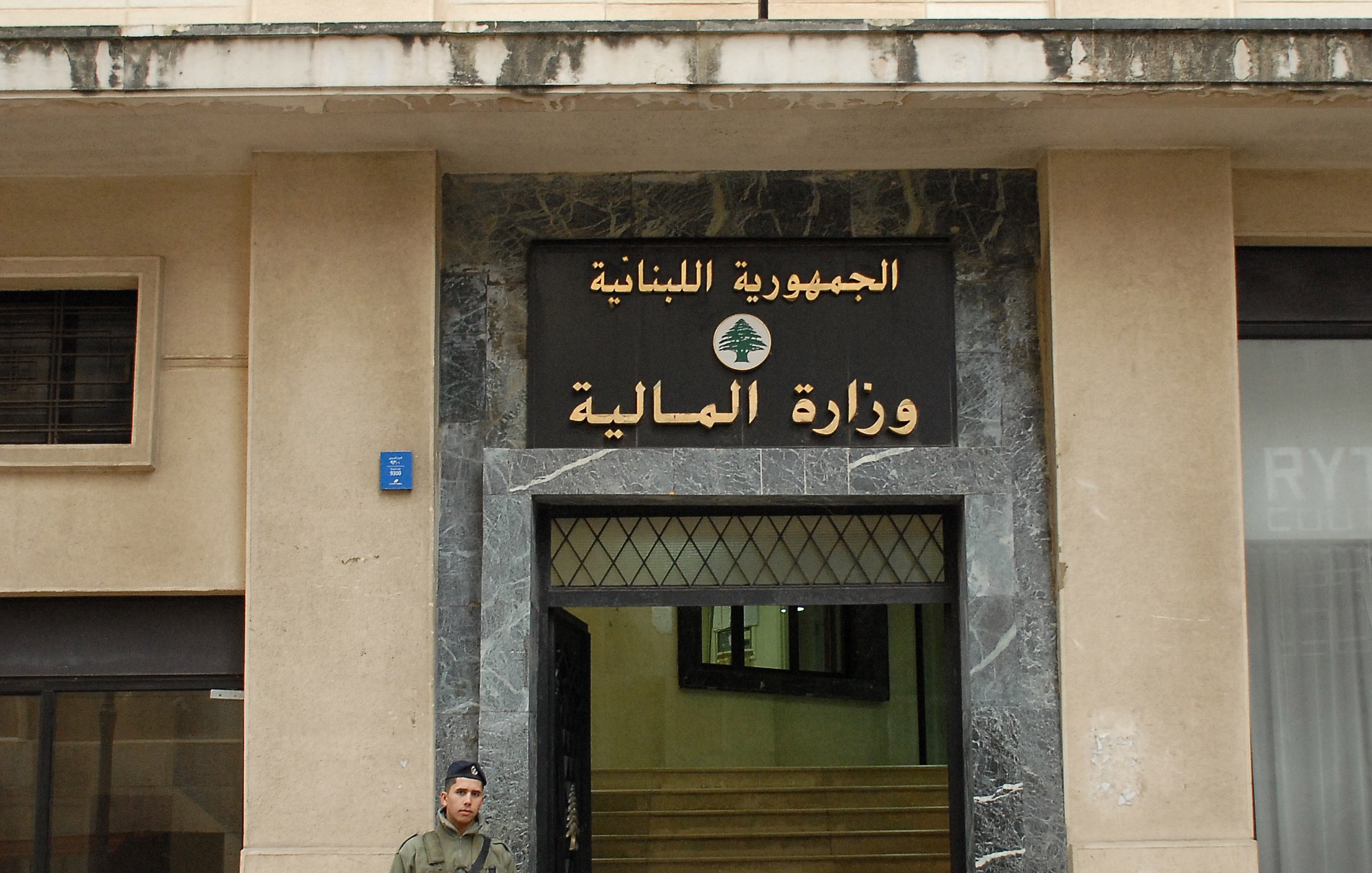 الصورة تظهر واجهة وزارة المالية في لبنان، والتي تحمل العلامة الرسمية للجمهورية اللبنانية. يوجد على الجهة اليسرى من الصورة جندي يقف حارسًا، يحمل سلاحًا، وقد ارتدى زيًا عسكريًا. المبنى يبدو بسيطًا وعصريًا، ويعكس الطراز المعماري الحضري.