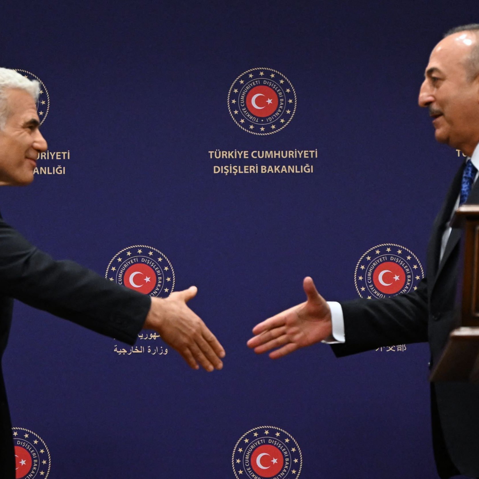 في الصورة، يظهر اثنان من الرجال يقفان في مواجهة بعضهما البعض. يبدو أن أحدهما يقدم يده لمصافحة الآخر. خلفهما يظهر علم تركيا مع شعار وزارة الخارجية التركية. الأجواء تبدو رسمية، مما يشير إلى أن اللقاء قد يكون متعلقاً بموضوع دبلوماسي أو رسمي بين البلدين.