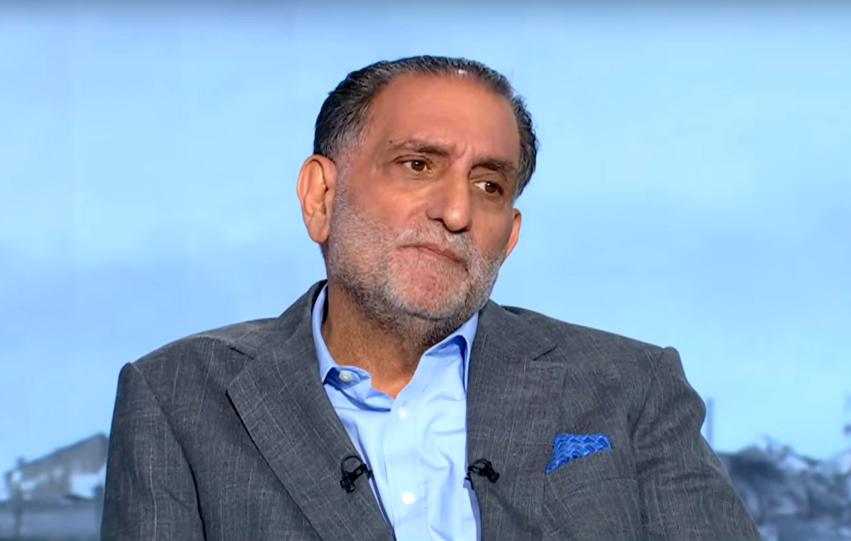 Homme en costume gris, assis, avec une expression pensante, arrière-plan flou.