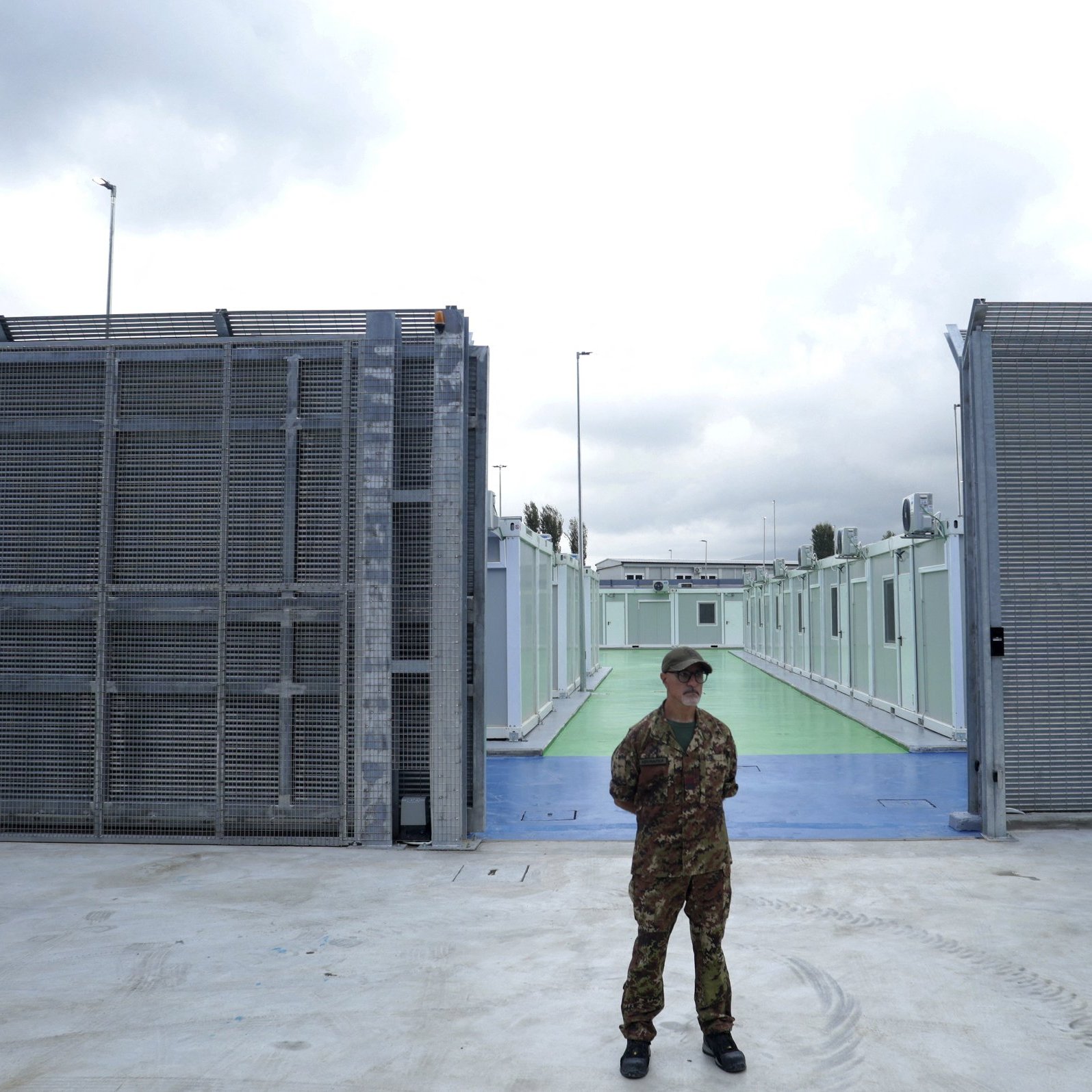L'immagine mostra un soldato in uniforme, in piedi al centro di un'area delimitata. Sullo sfondo si possono vedere due strutture metalliche grigie che sembrano essere recinzioni, con un'area di pavimentazione colorata di verde e blu tra di esse. Il cielo è nuvoloso, suggerendo una giornata grigia. L'atmosfera sembra essere di sicurezza e controllo, con un forte senso di vigilanza da parte del soldato.
