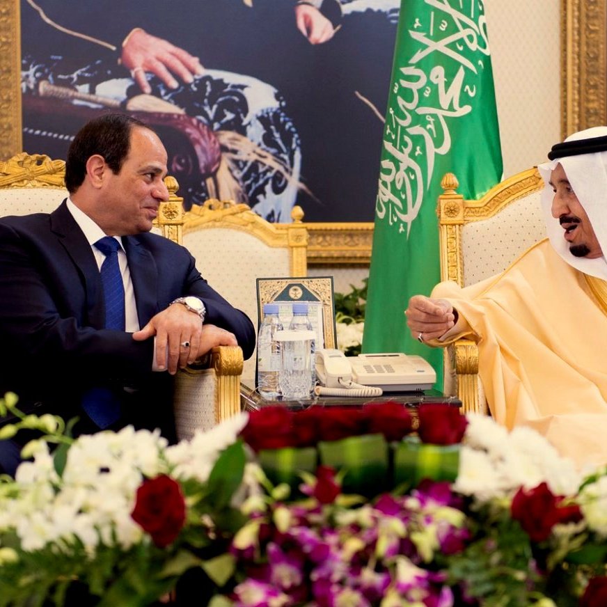 L'image montre deux hommes assis face à face dans un cadre officiel. Ils sont vêtus de manière formelle : l'un porte un costume noir avec une cravate, tandis que l'autre est en tenue traditionnelle dorée. Ils sont entourés d'une décoration luxueuse, avec des drapeaux en arrière-plan et des fleurs sur une table basse. L'atmosphère semble conviviale et sérieuse à la fois, suggérant une discussion importante.