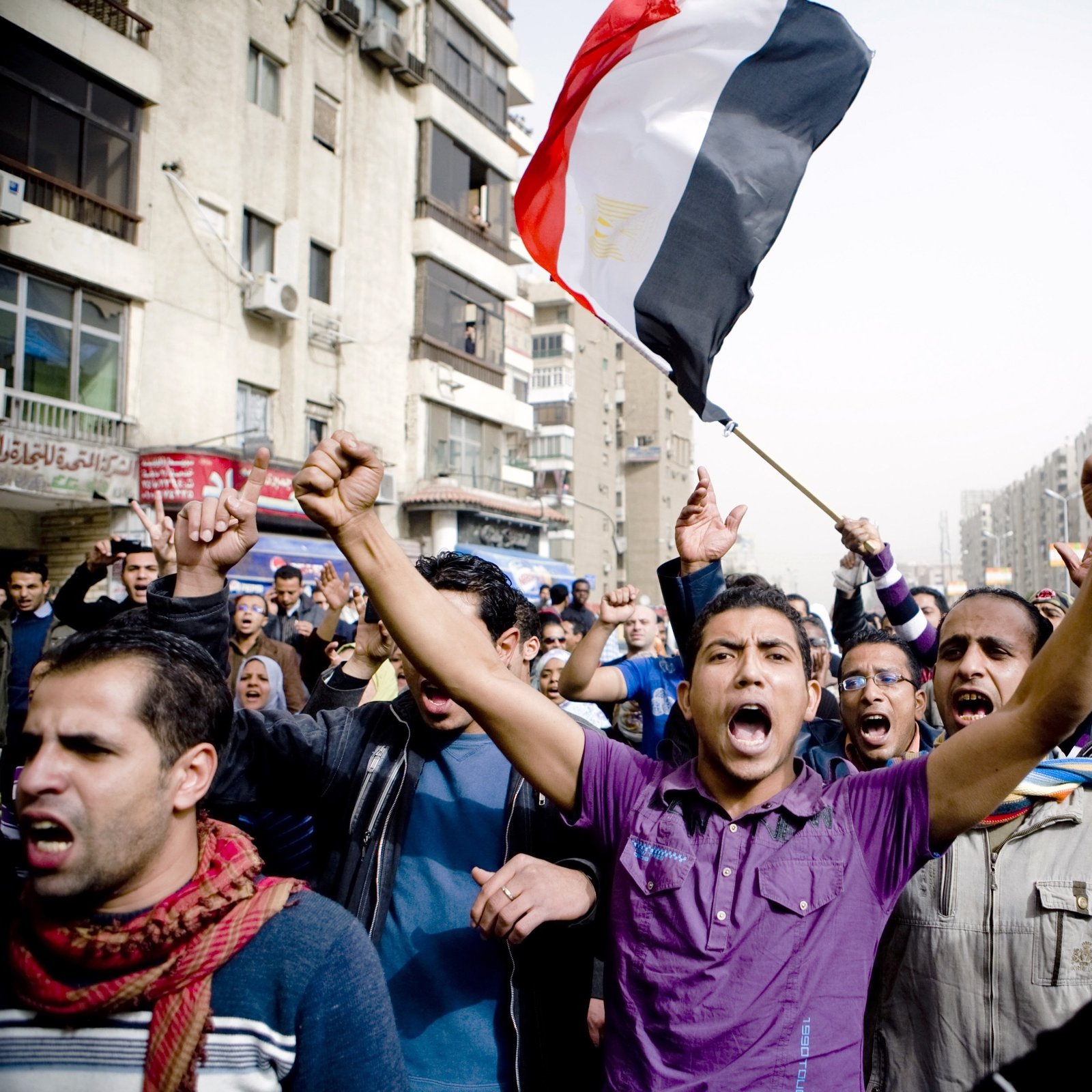 الصورة تظهر مجموعة من الناس في مظاهرة حاشدة. في المقدمة، يوجد رجل يحمل علم مصر ويبدو أنه يرفع صوته عالياً تعبيراً عن الحماس أو المطالب. خلفه يظهر حشد كبير من المتظاهرين، بعضهم يرفعون أيديهم ويهتفون. تعكس الصورة أجواء من النشاط والمشاركة الشعبية، مع مباني المدينة في الخلفية تحت سماء واضحة.