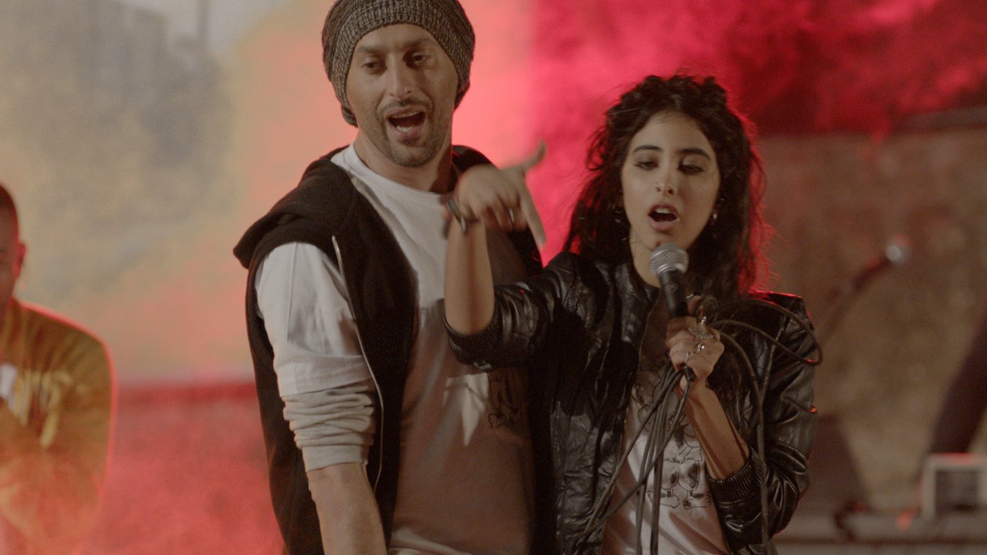 L'image montre des artistes en pleine performance musicale, avec une ambiance vibrante et colorée. Au centre, une femme chante dans un microphone, accompagnée d'un homme à ses côtés qui semble participer activement à la performance. En arrière-plan, d'autres musiciens se joignent à l'animation, entourés de lumière et de fumée, créant une atmosphère dynamique et festive. Les expressions des artistes indiquent de l'enthousiasme et de l'énergie.