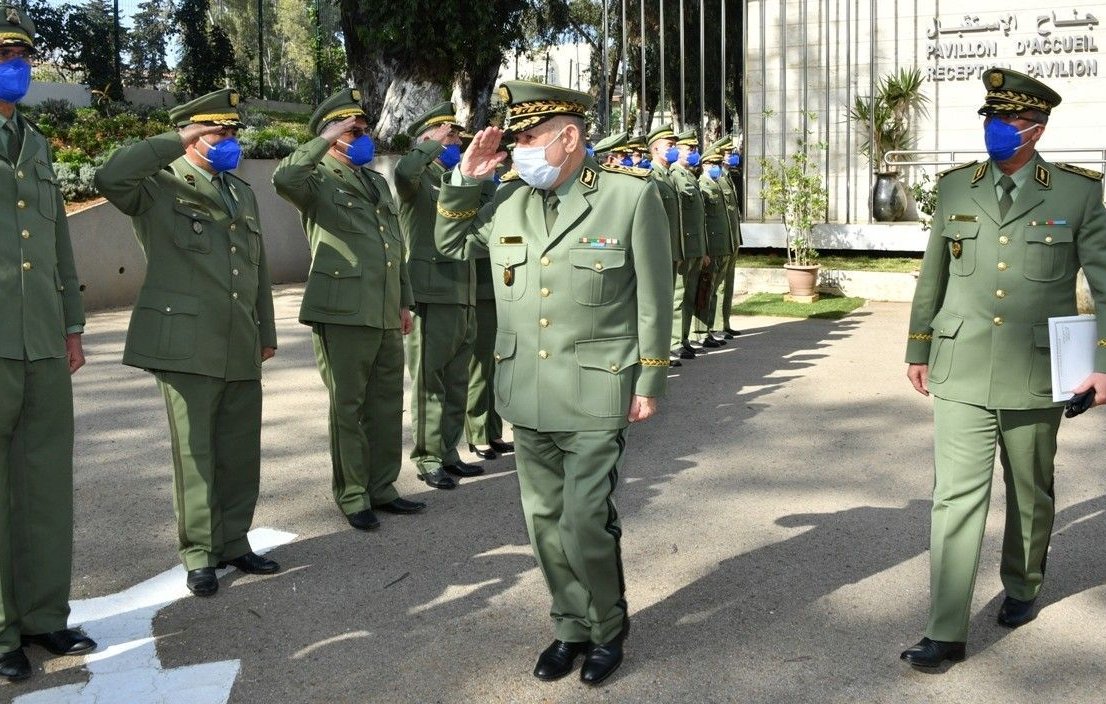 L'image montre un groupe de militaires en uniforme, saluant un supérieur. Tous portent des masques et des casquettes, ce qui indique qu'ils respectent des protocoles de santé. L'environnement semble être un site officiel ou militaire, avec des bâtiments en arrière-plan. Les militaires adoptent une posture de respect, ce qui suggère une cérémonie ou un événement formel.