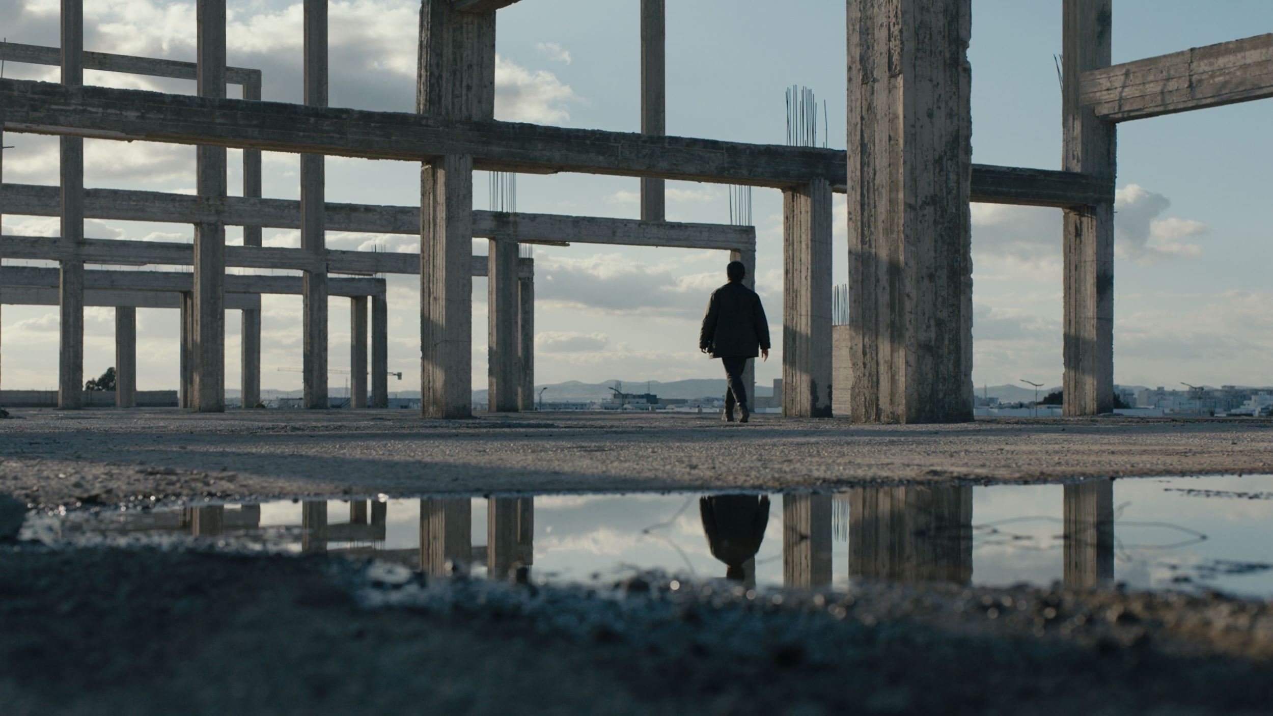 L'image montre une personne marchant à travers une structure de bâtiment inachevée. Les piliers en béton se dressent autour d'elle, créant une atmosphère à la fois vide et mystérieuse. Le sol est humide, et une flaque d'eau reflète les piliers et le ciel, ajoutant une dimension visuelle intéressante à la scène. Le ciel est partiellement nuageux, et l'éclairage est doux, contribuant à l'ambiance contemplative de cet espace abandonné.